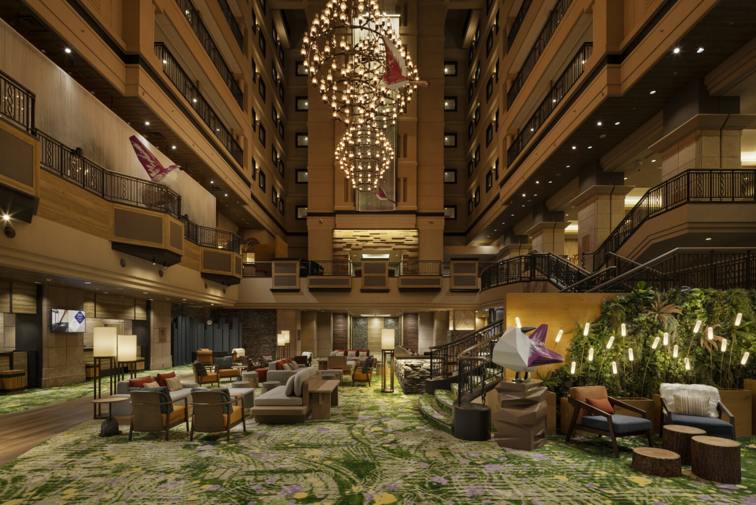 Spacious hotel lobby with green-patterned carpet, warm lighting, and modern seating. A grand chandelier and decorative greenery create a welcoming atmosphere.