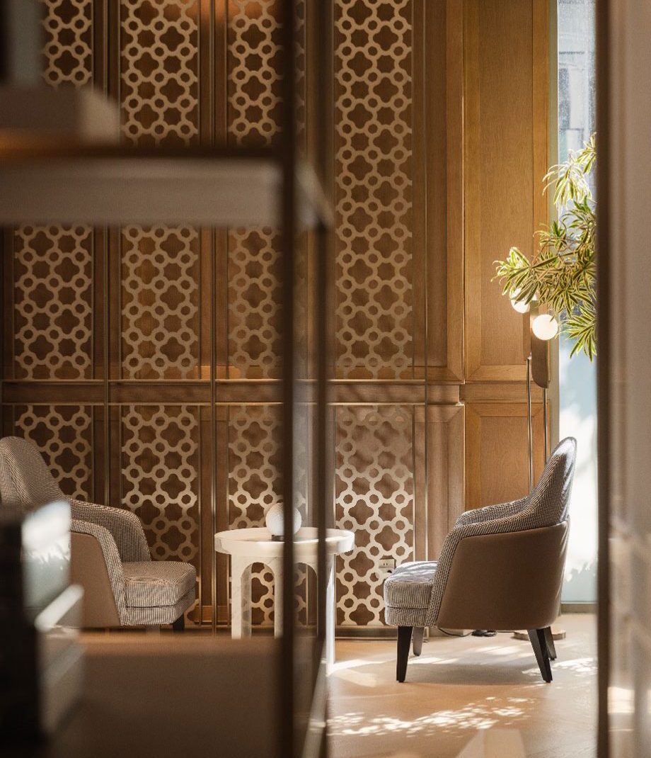 Elegant lounge with two patterned armchairs facing a small round table. A geometric wooden panel and a potted plant create a warm, inviting atmosphere.