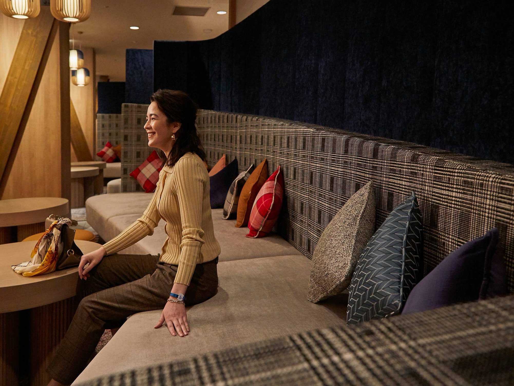 A smiling woman sits on a plaid couch with colorful pillows in a modern, warmly lit lounge. The atmosphere is cozy and relaxed.