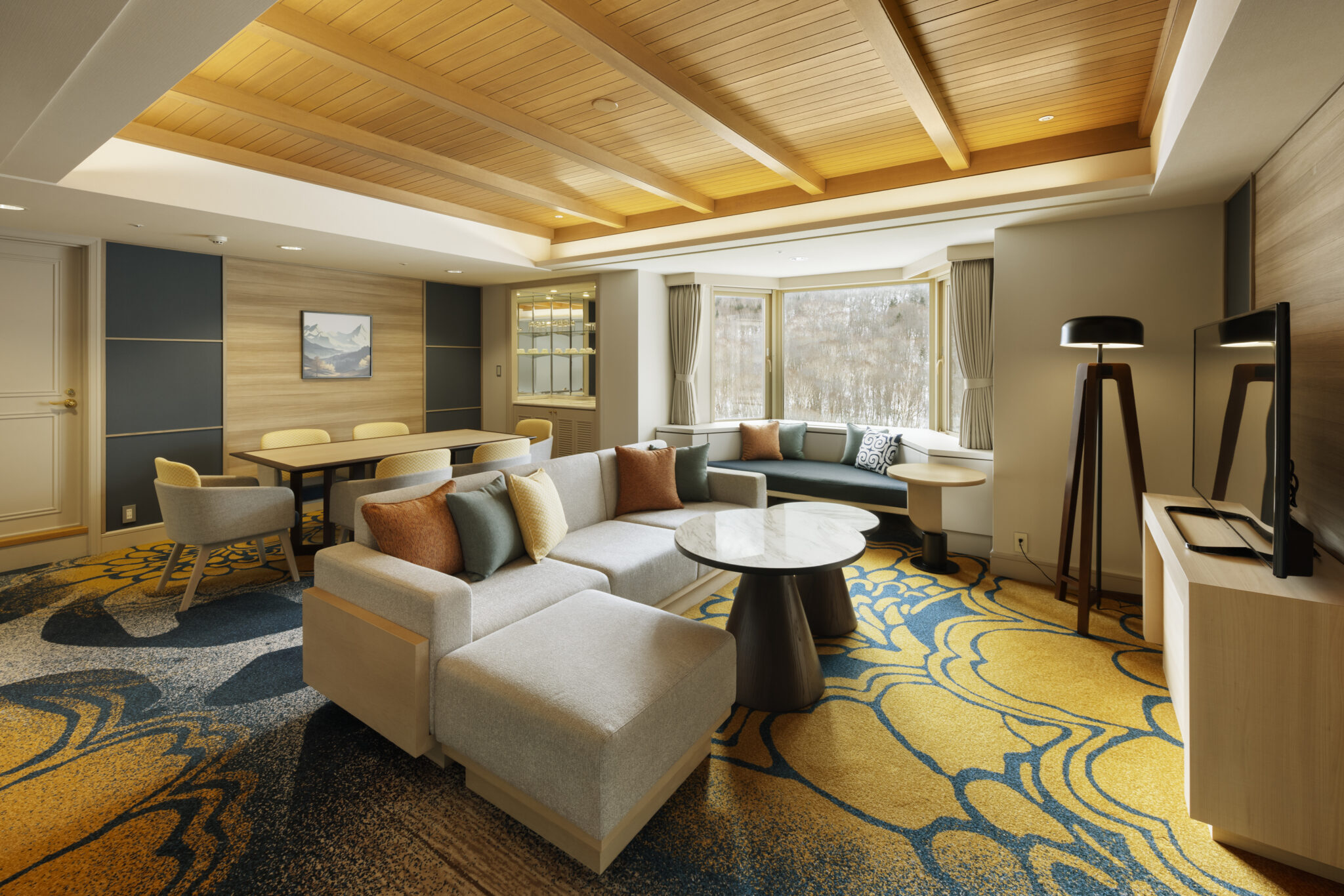 Spacious living area in hotel's family suite with cozy gray sofas, colorful cushions, vibrant yellow and blue carpet, wooden ceiling, large window, and modern decor.