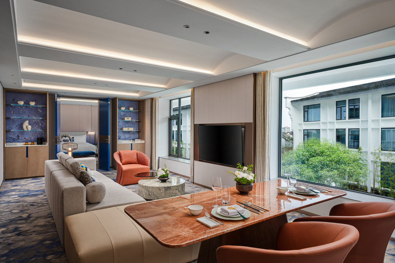 Modern hotel suite with sleek furniture, a cozy orange chair, large windows showing greenery, and a dining area with set tableware, exuding elegance.