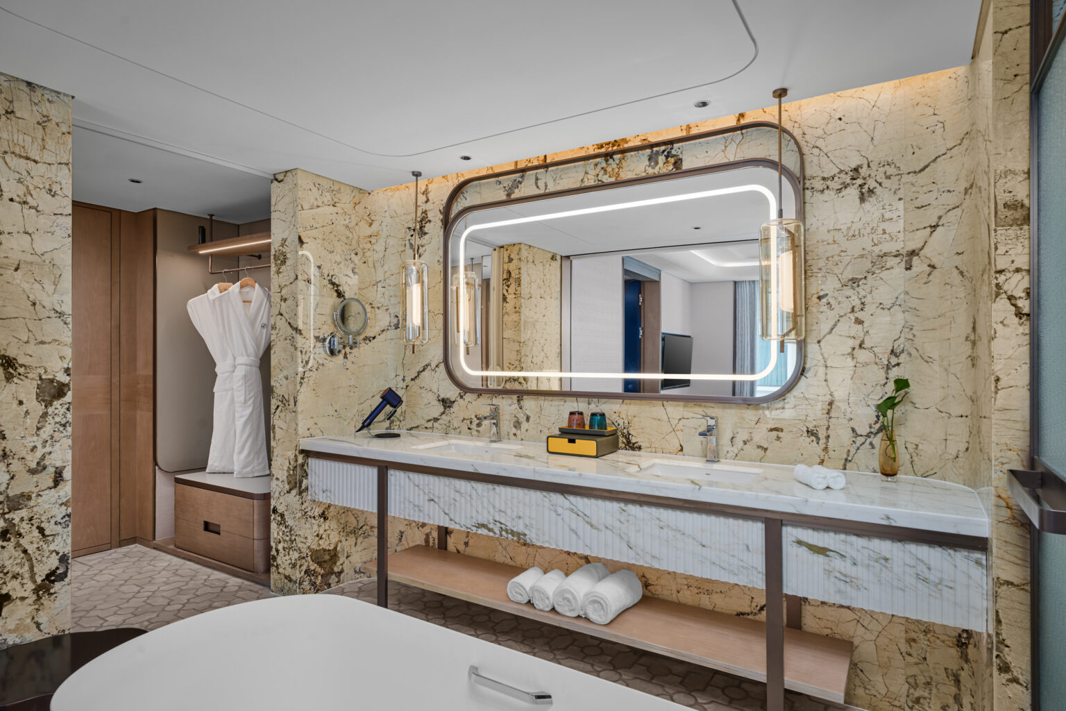 Luxurious bathroom with marbled walls, large illuminated mirror, and double sink vanity. A minimalist closet with bathrobes and an elegant bathtub enhance the spa-like ambiance.