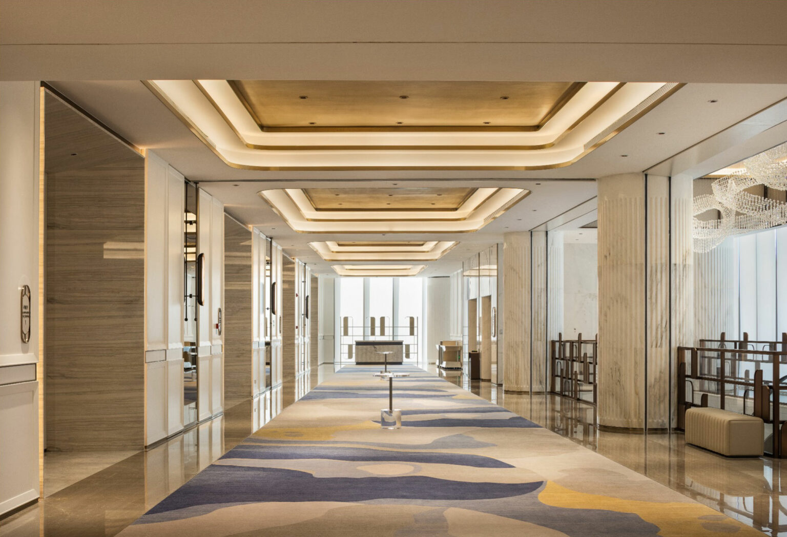 Luxurious prefunction area with elegant lighting and mirrored walls. The modern design features a patterned carpet in blue, beige, and yellow, creating a serene ambiance.