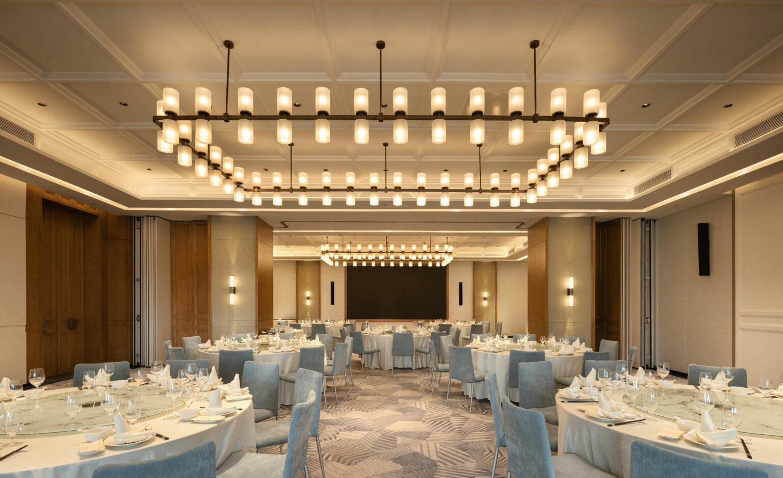 Elegant banquet hall with round tables set for dining, featuring white tablecloths, blue chairs, and ornate chandeliers. The scene is warm and inviting.