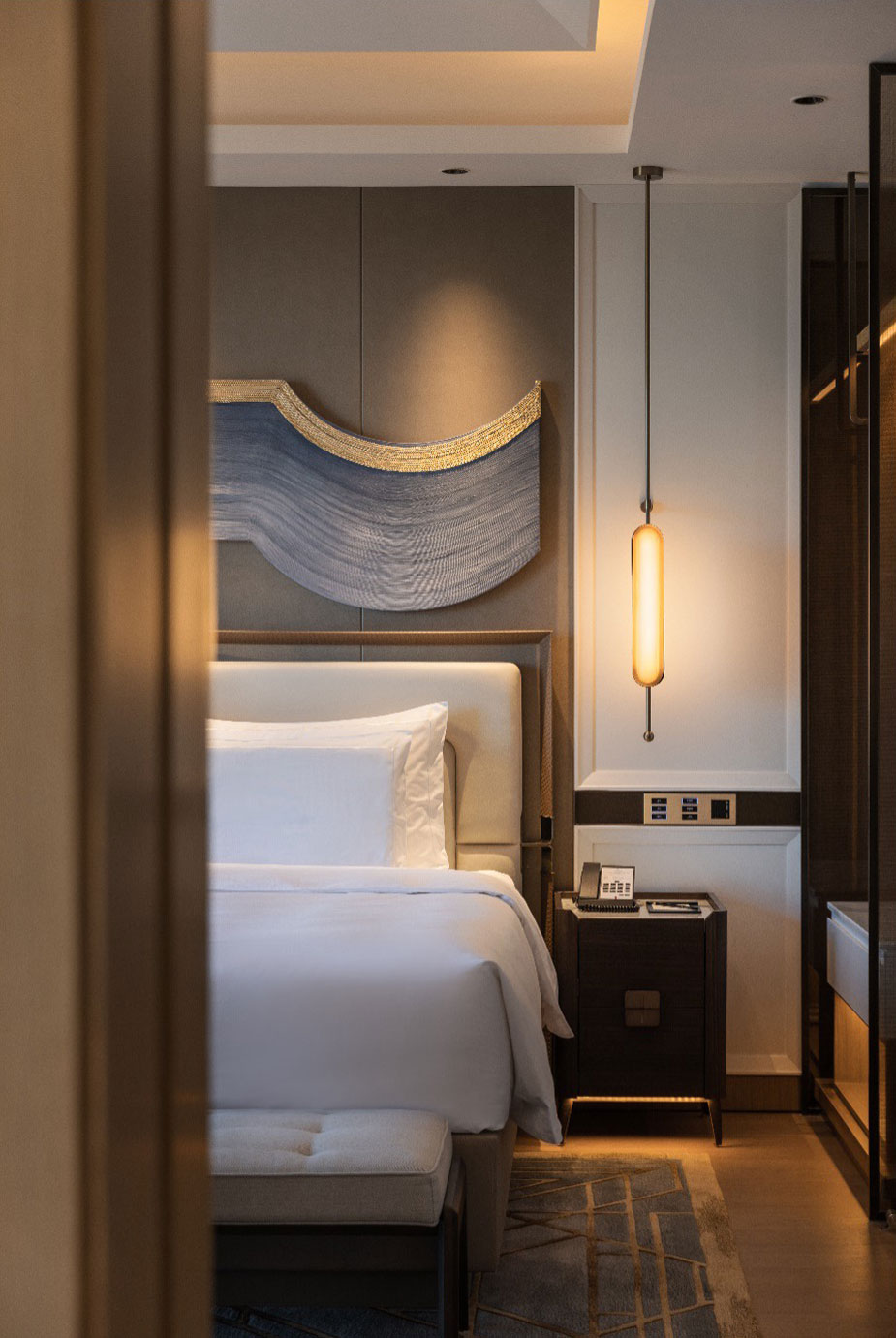 Modern hotel room with a cozy bed and white linens, featuring a warm glow from a pendant light. Abstract wall art adds elegance above the bed.