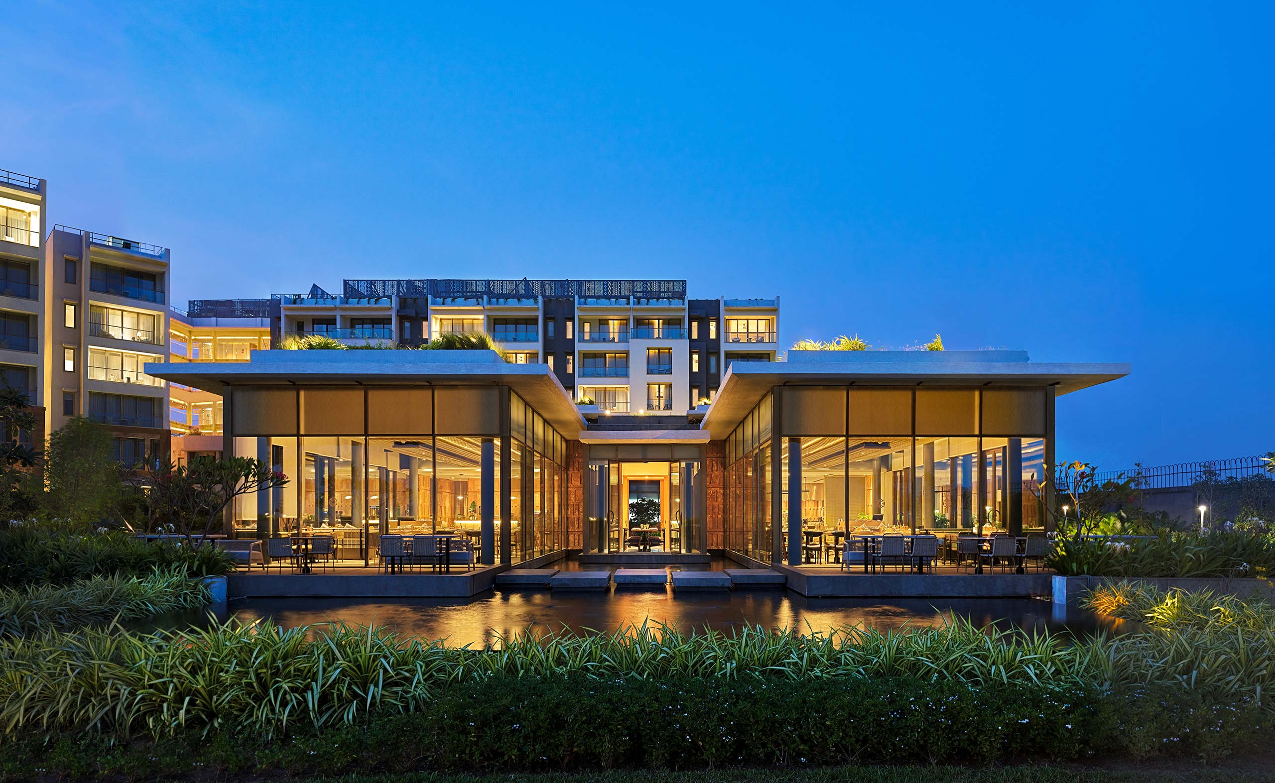 A modern building at dusk features large glass windows glowing warmly, surrounded by lush greenery and reflecting in a tranquil pond, exuding elegance and serenity.
