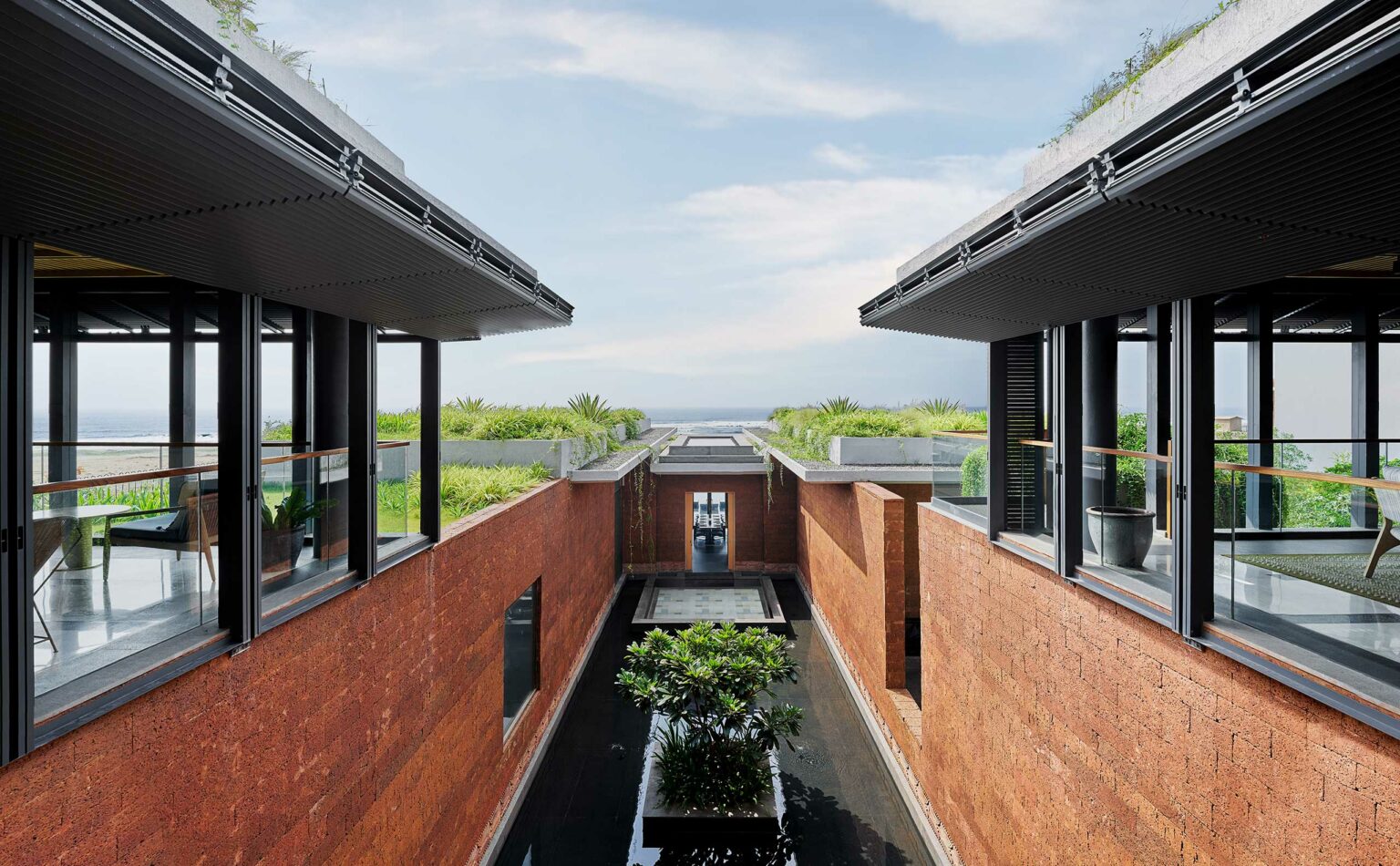 Modern architecture featuring parallel brick buildings with large windows, surrounding a central water feature with lush greenery, under a clear sky.