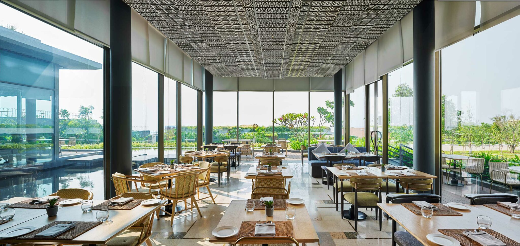 Spacious, modern restaurant with large windows showcasing a lush garden view. Wooden tables, chairs, and soft lighting create a serene dining ambiance.