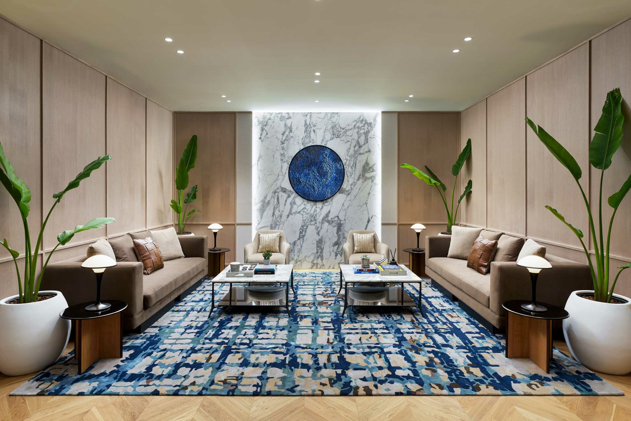 Modern living room with two beige sofas facing each other, separated by two white coffee tables on a blue patterned rug. White walls feature large plants and a central marble panel with a textured blue circle. Bright, elegant ambiance.