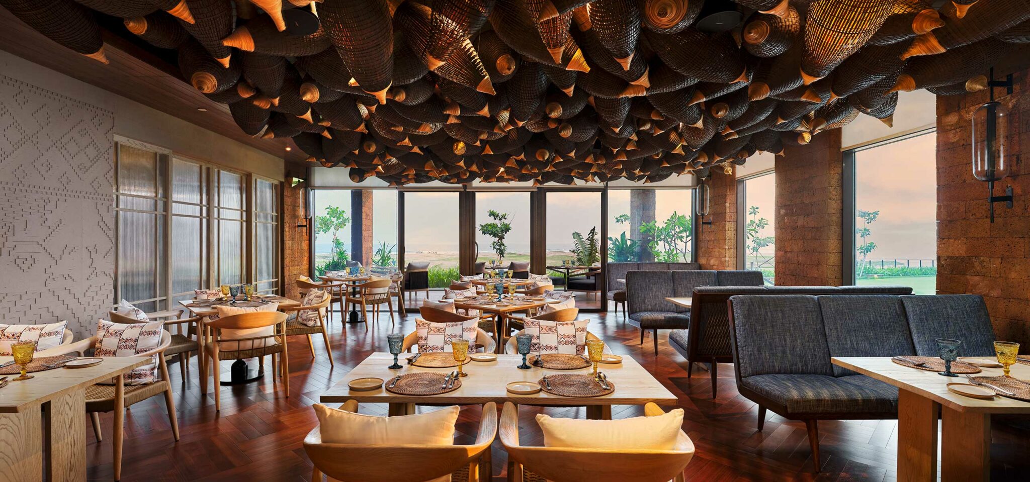 Elegant restaurant interior with a warm, sophisticated vibe. Features woven decor on the ceiling, wooden floors, modern tables, cushioned chairs, and large windows with a scenic view.