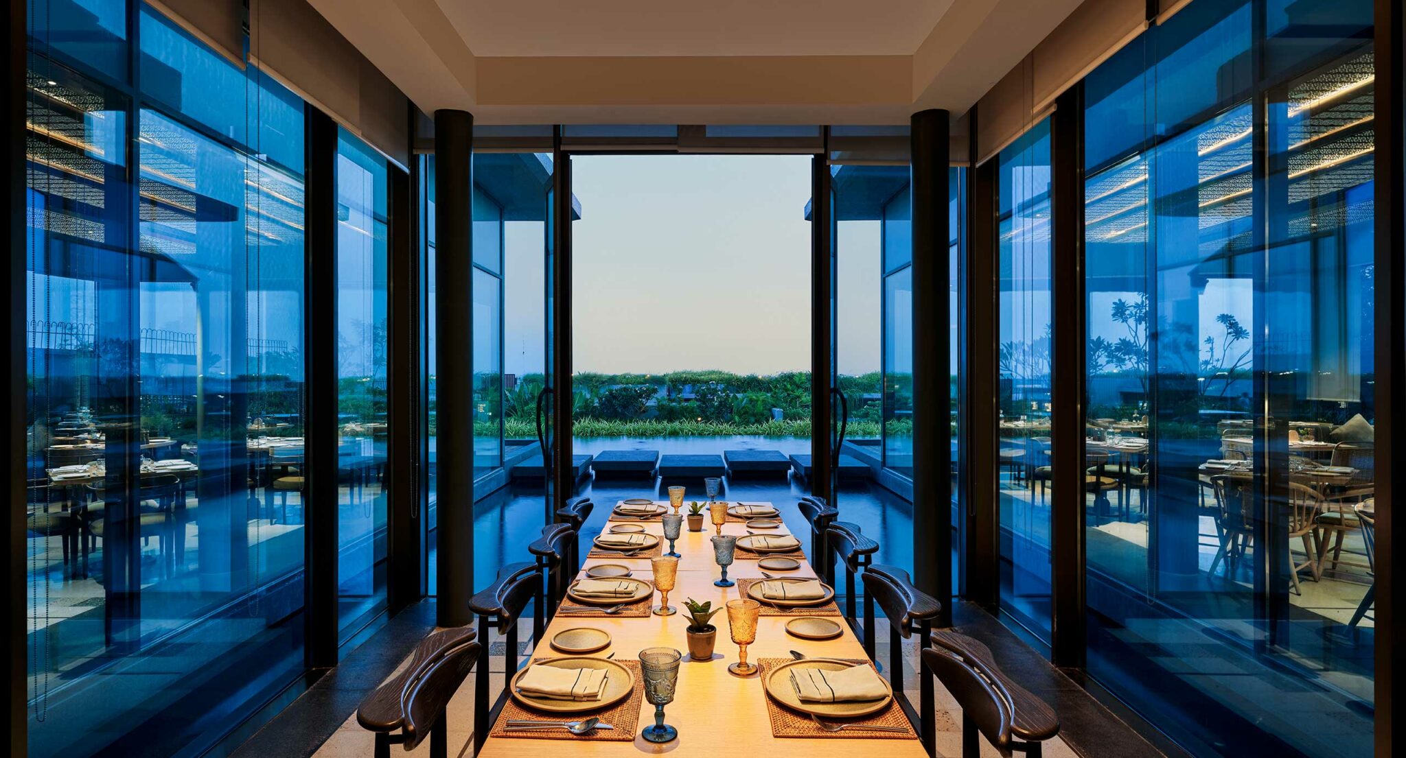 Elegant dining room with a long wooden table set for eight, surrounded by reflective glass walls. A serene outdoor view is visible at dusk.