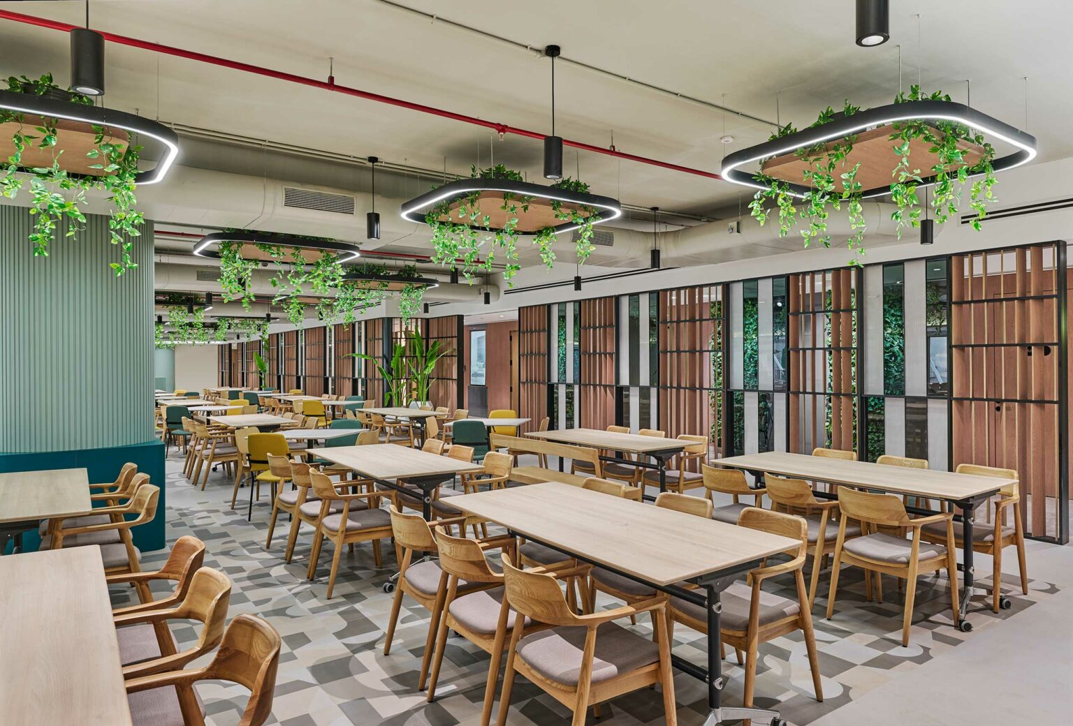 Spacious modern cafeteria with wooden tables and chairs, greenery hanging from ceiling light fixtures, and stylish vertical wood paneling on walls.