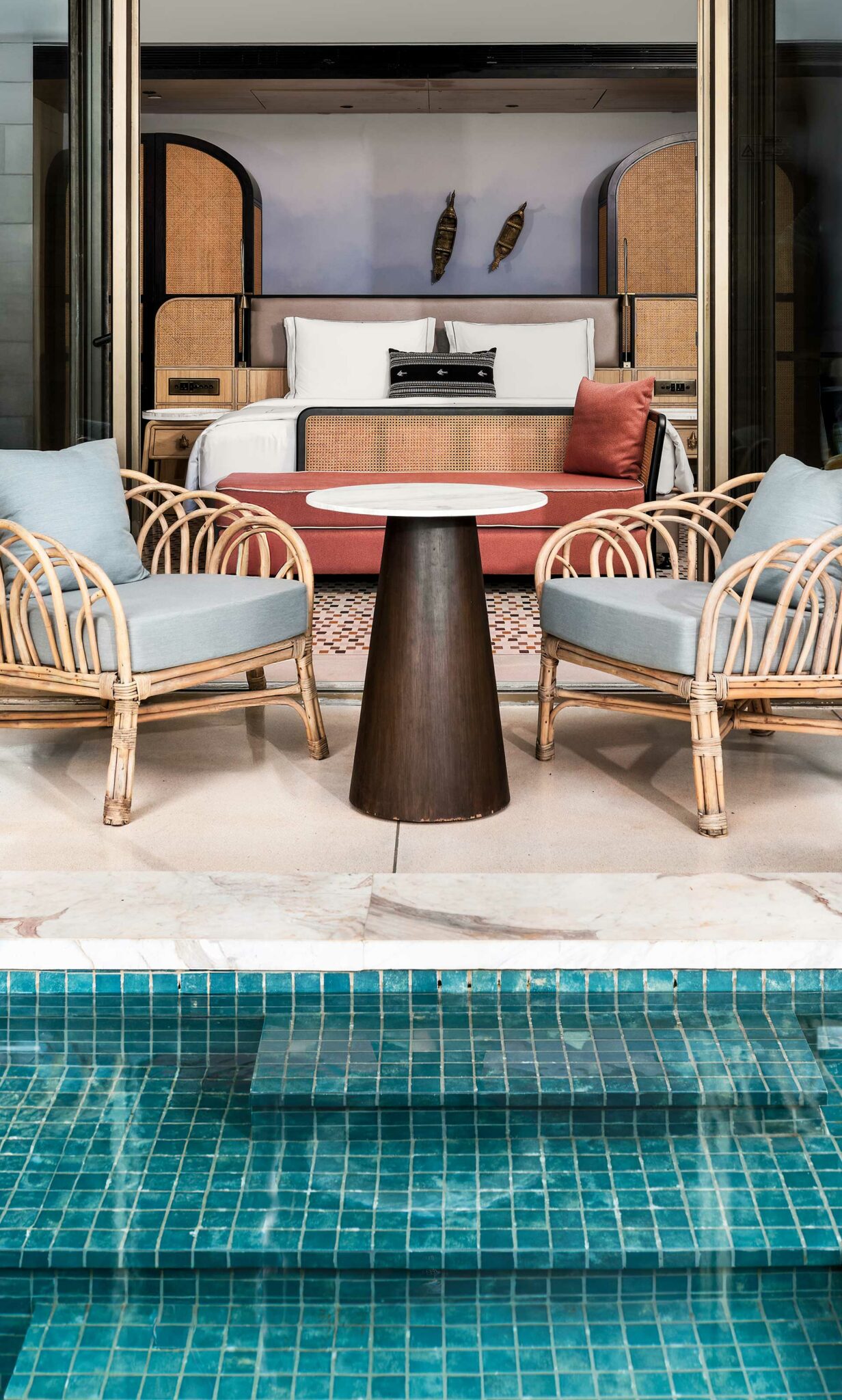 A cozy poolside setup with two rattan chairs, light blue cushions, and a small round table on a patio. Behind is a stylish bed with wooden accents.