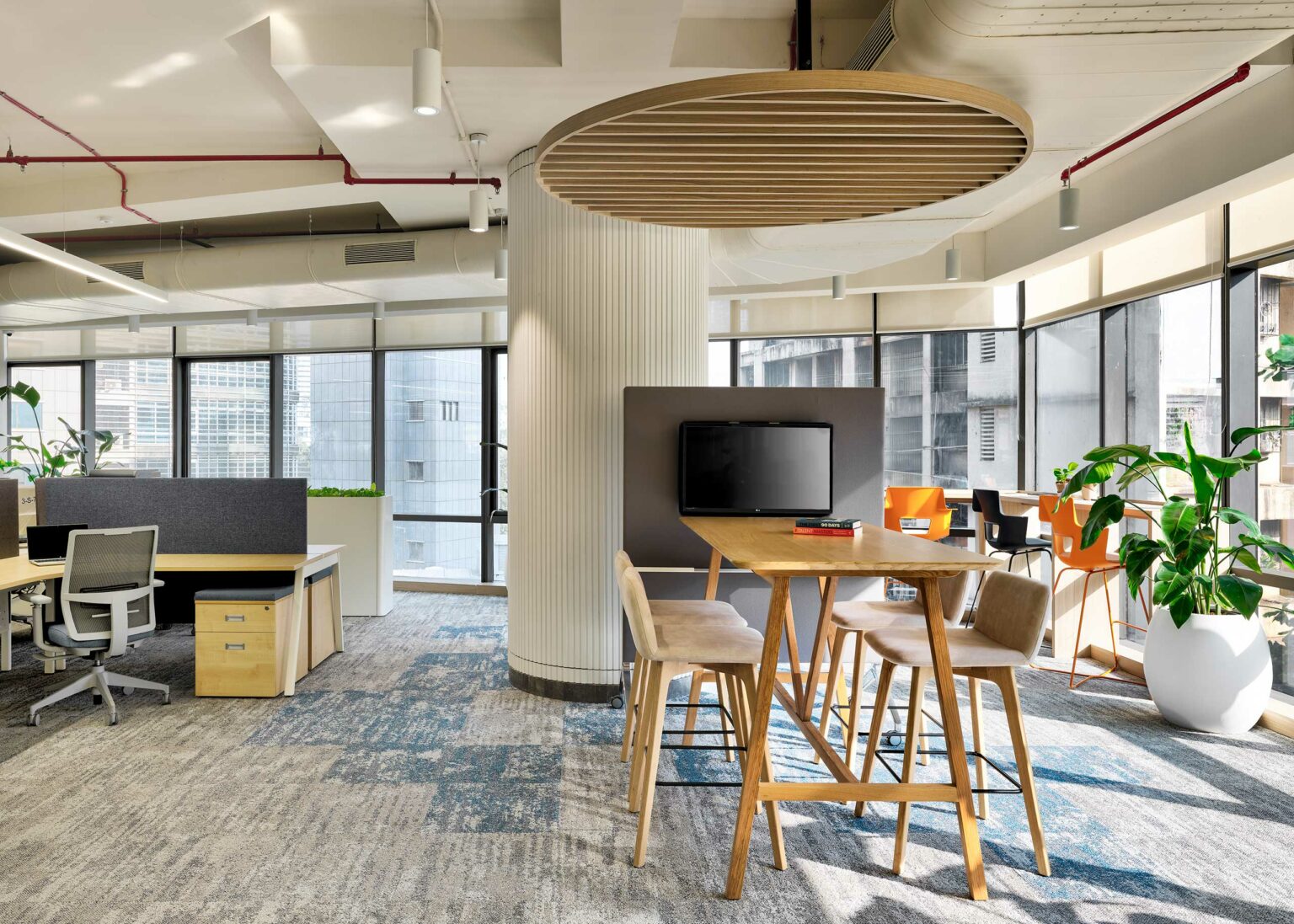 Modern office space with high ceilings, featuring a wooden meeting table with four chairs in the foreground. Large windows, plants, and a casual, airy ambiance.