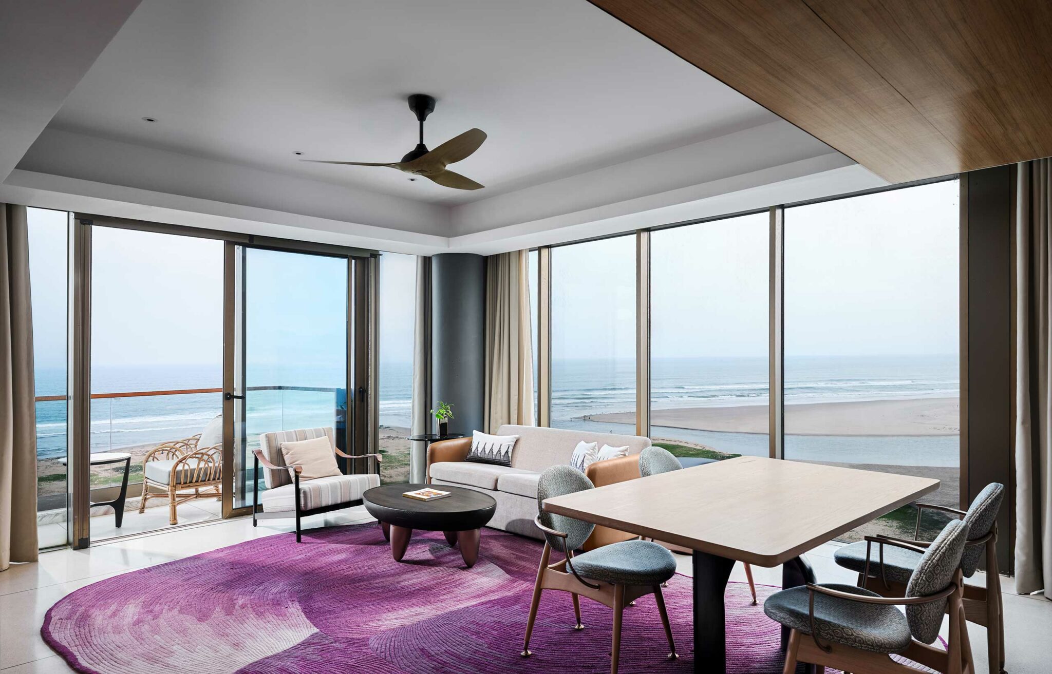Modern living room with ocean view, featuring large windows, a beige sofa, wooden table, chairs, and a purple rug, creating a tranquil atmosphere.