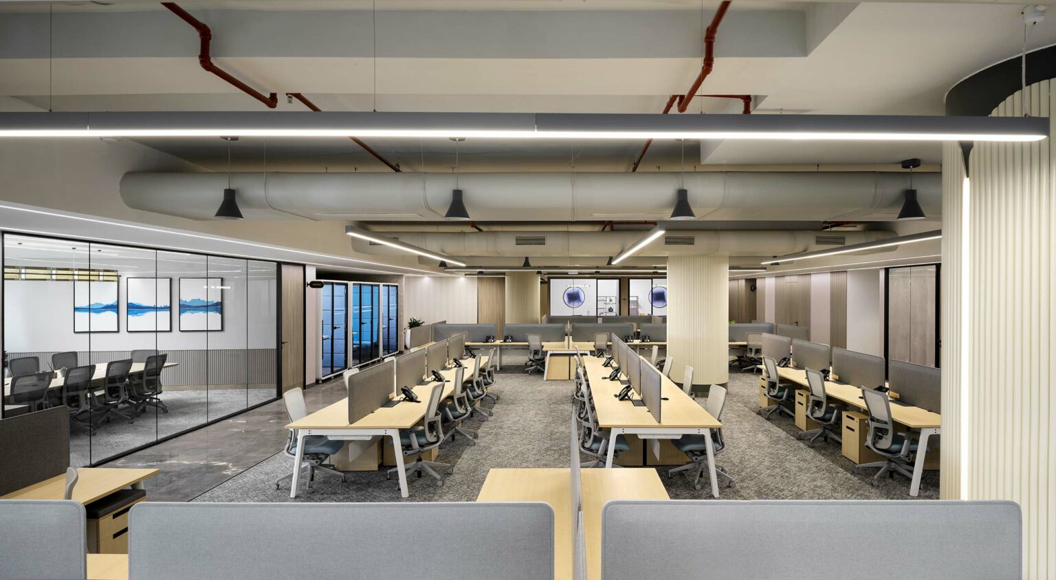 Modern open-plan office with rows of desks and grey privacy dividers. Soft lighting, exposed pipes, and glass-walled meeting rooms create a sleek, professional atmosphere.