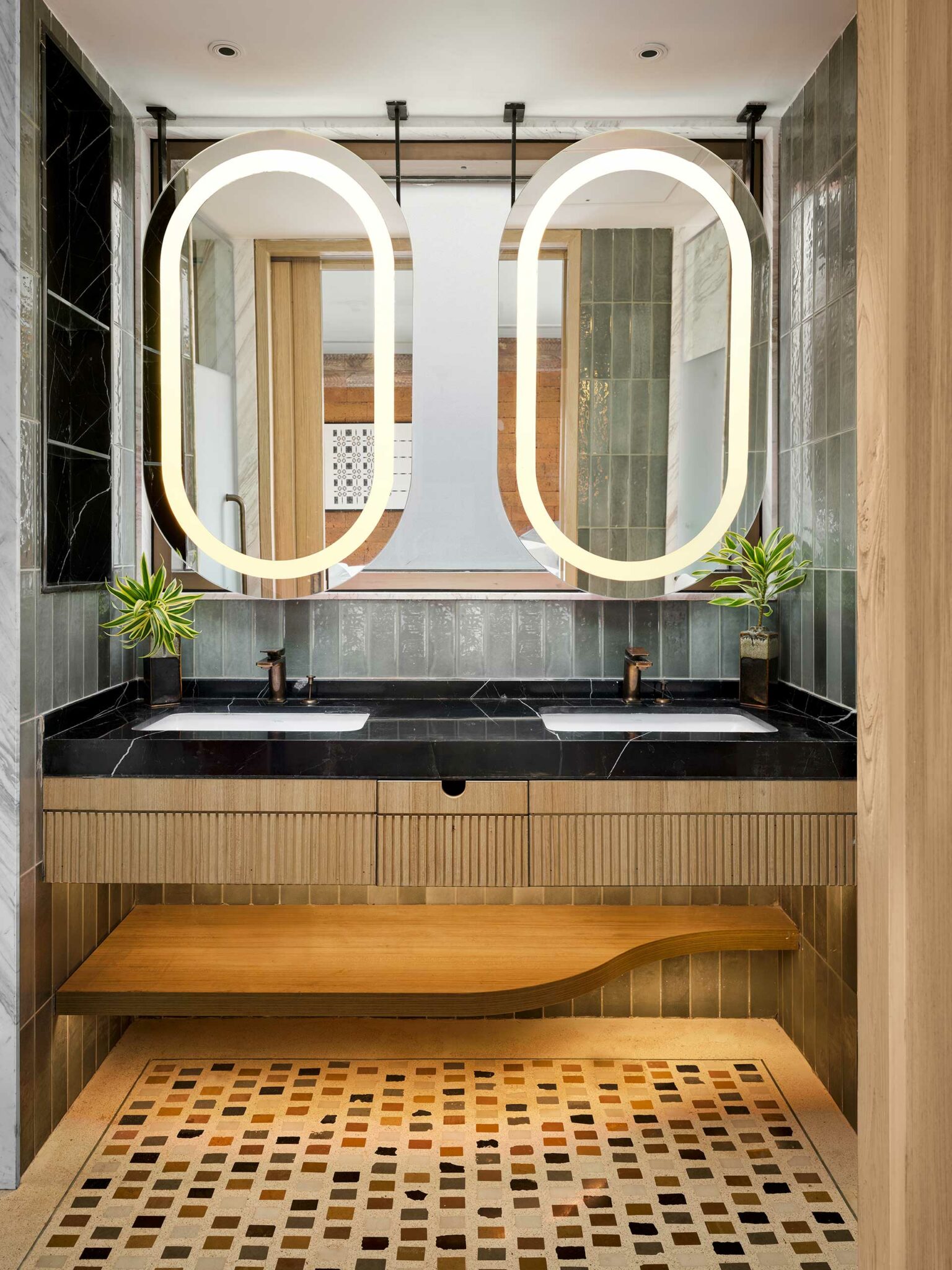 Modern bathroom with dual oval mirrors above a black countertop and double sinks. The decor includes green plants and elegant lighting, creating a sleek, tranquil ambiance.