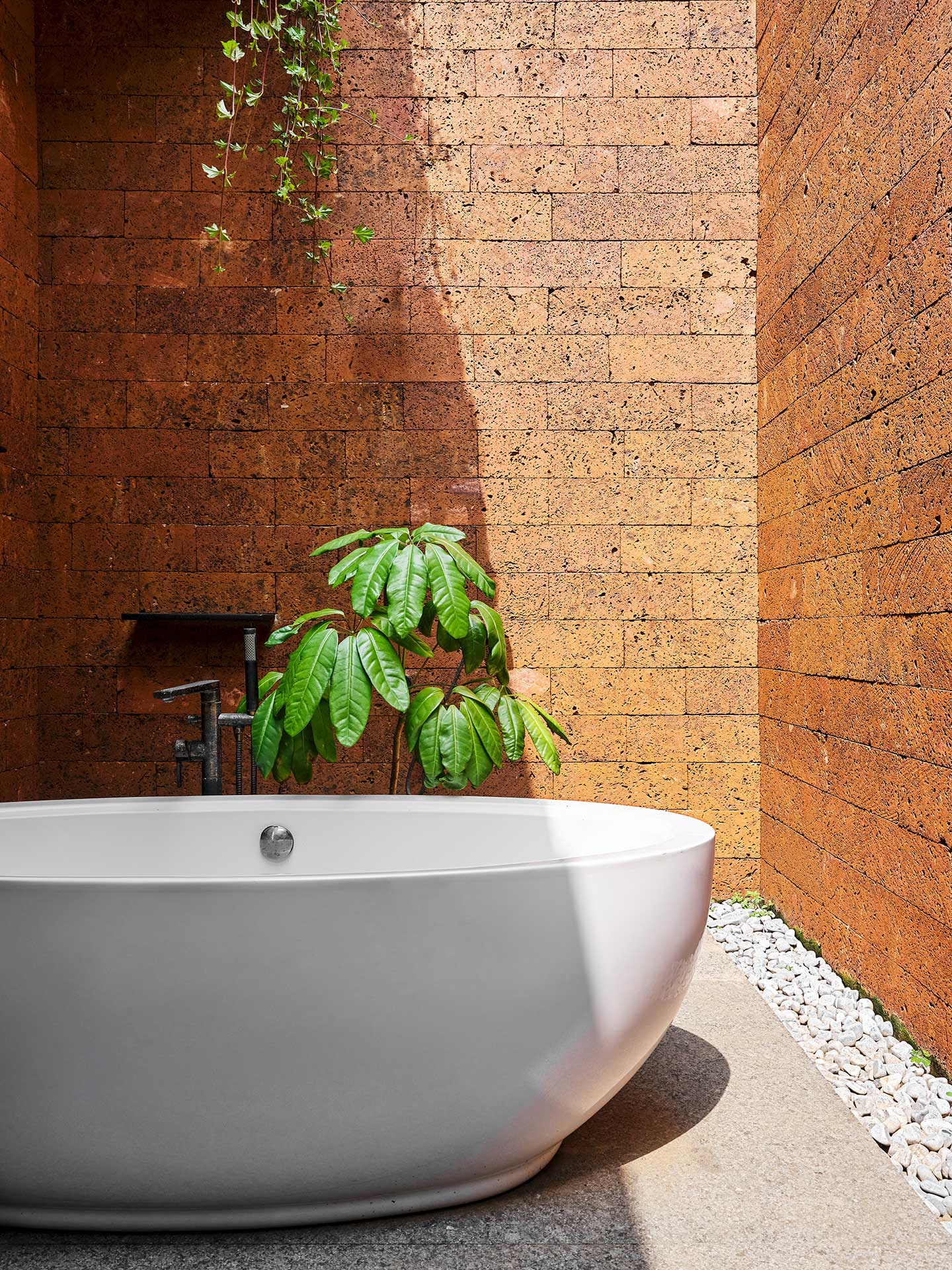 Minimalist outdoor bath scene with a round white tub against a textured orange brick wall, lush green plant, and cascade of vines, creating a serene ambiance.