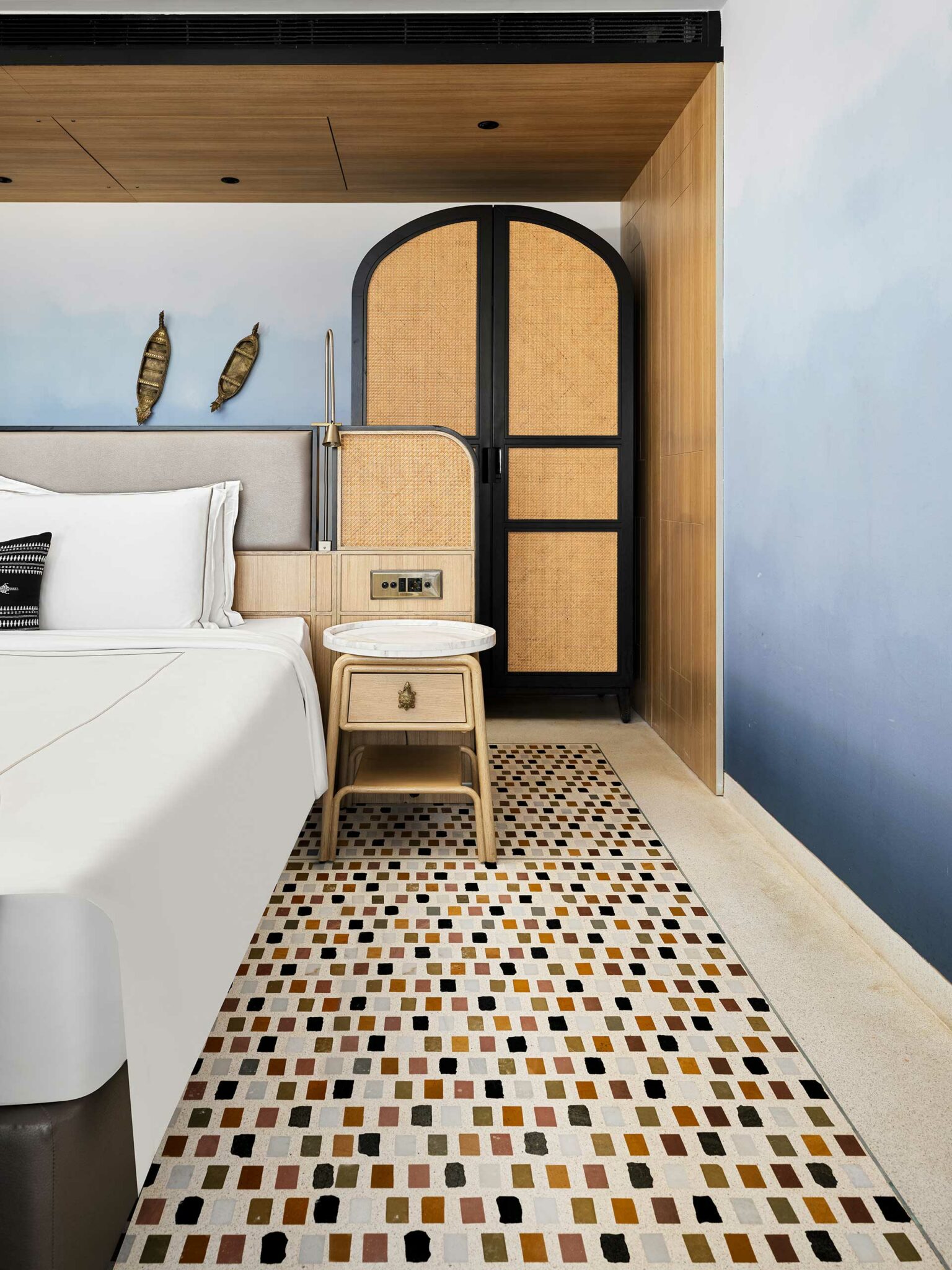 Stylish hotel room with a white bed, a wooden side table, and a woven wardrobe. The floor has a colorful mosaic carpet, evoking a cozy, modern feel.