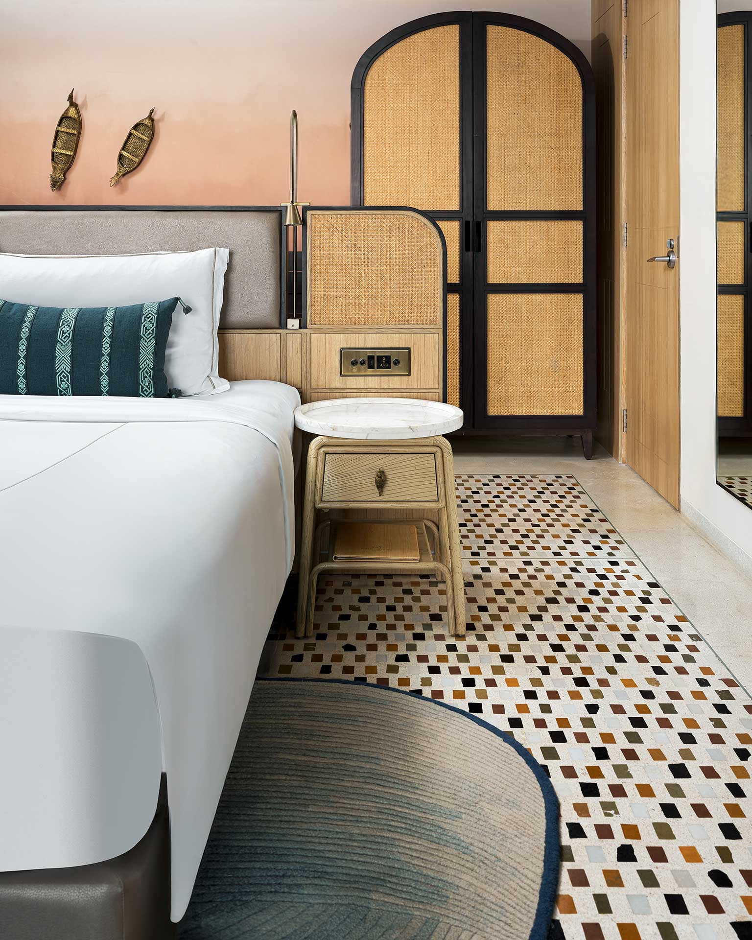 Modern bedroom with a stylish white bed, teal cushion, rattan wardrobe, and woven wall decor. Warm tones, geometric rug, and cozy ambiance.