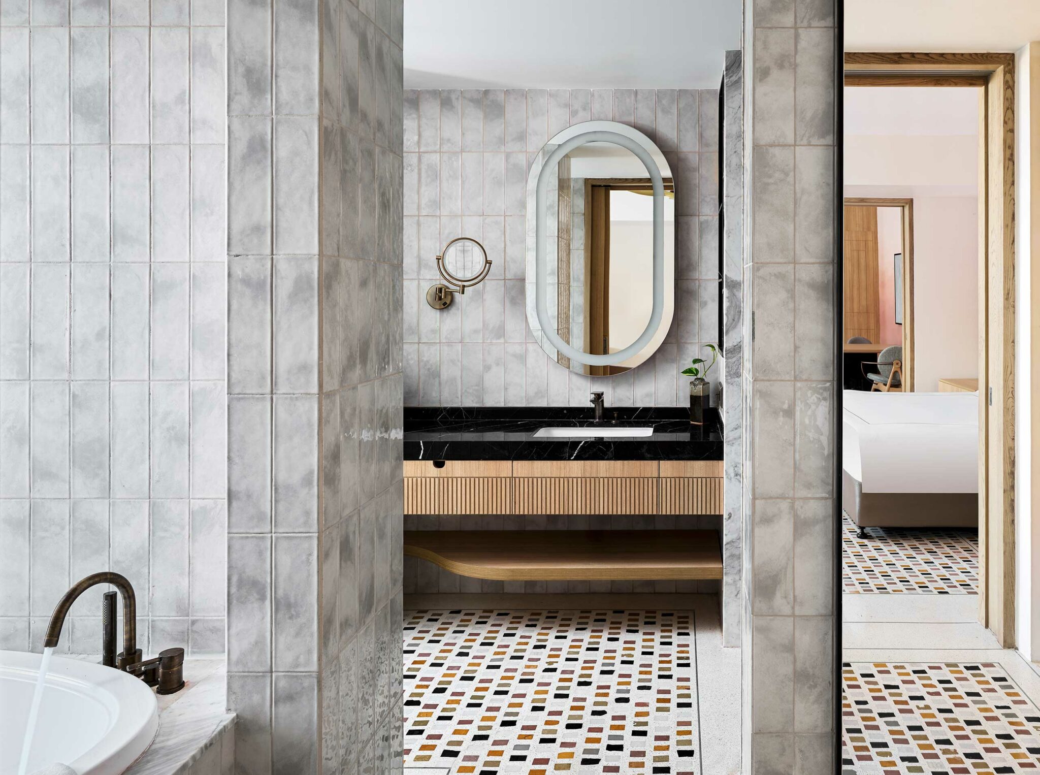 Modern bathroom with gray tiled walls and mosaic floor. Features a black countertop with an oval mirror, round magnifying mirror, and visible adjoining bedroom. Clean and elegant design.