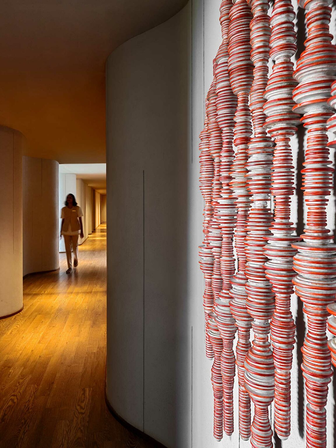 Curved hallway with wooden floor, featuring a wall with an artistic installation of stacked red and white discs in the foreground; a person walks in the background.
