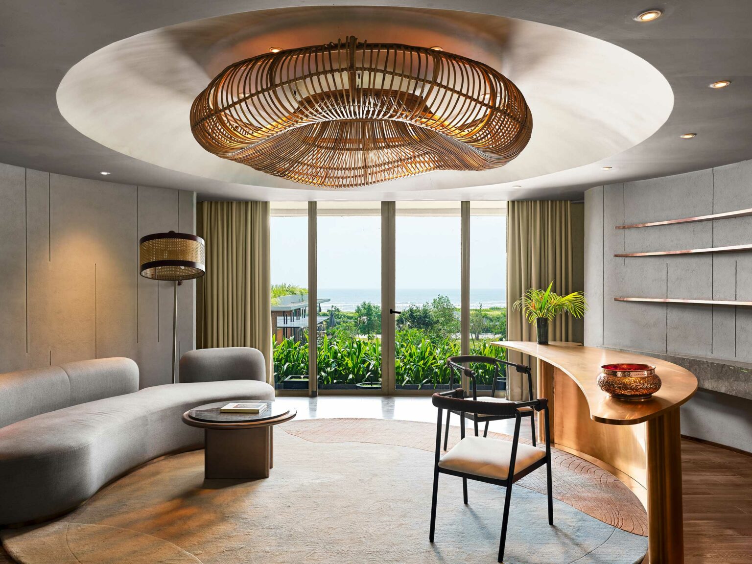 Modern Reception with a large wicker chandelier, curved gray sofa, and wooden table. Glass doors reveal lush greenery and ocean view, exuding tranquility.