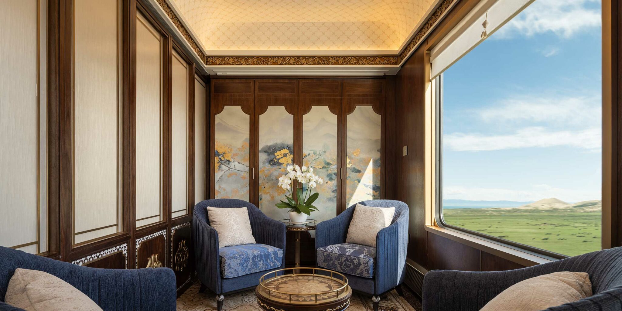 Luxurious train carriage interior with two blue armchairs facing a small round table. Large window reveals scenic landscape of green fields and blue sky.