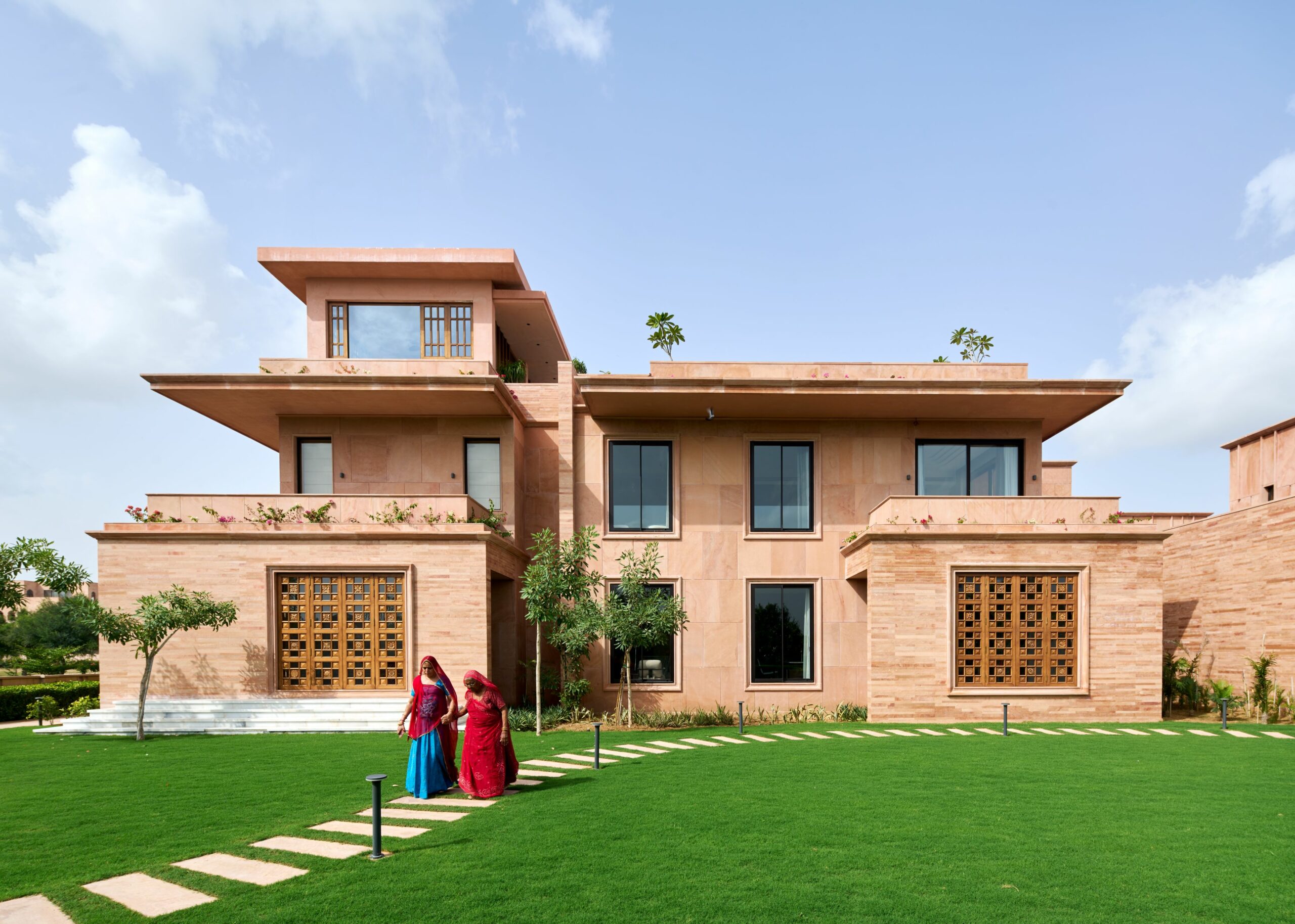 A large, modern house with a Sandstone facade and tall windows, set against a bright sky. Two people in colorful attire walk on a lush green lawn.