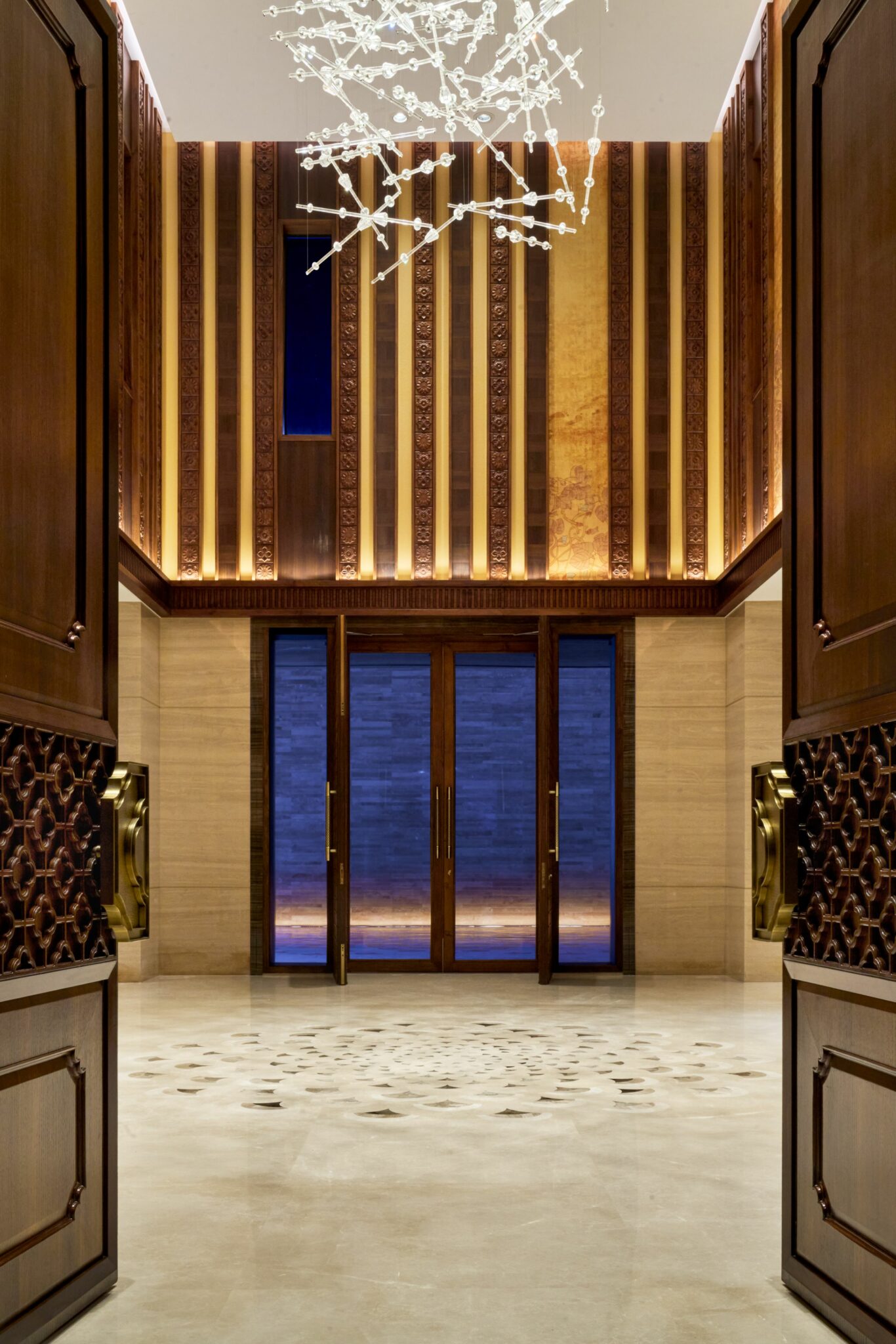 Luxurious foyer with tall wooden doors opening to a grand space. Intricate ceiling lights and elegant wood paneling create a warm, opulent atmosphere.