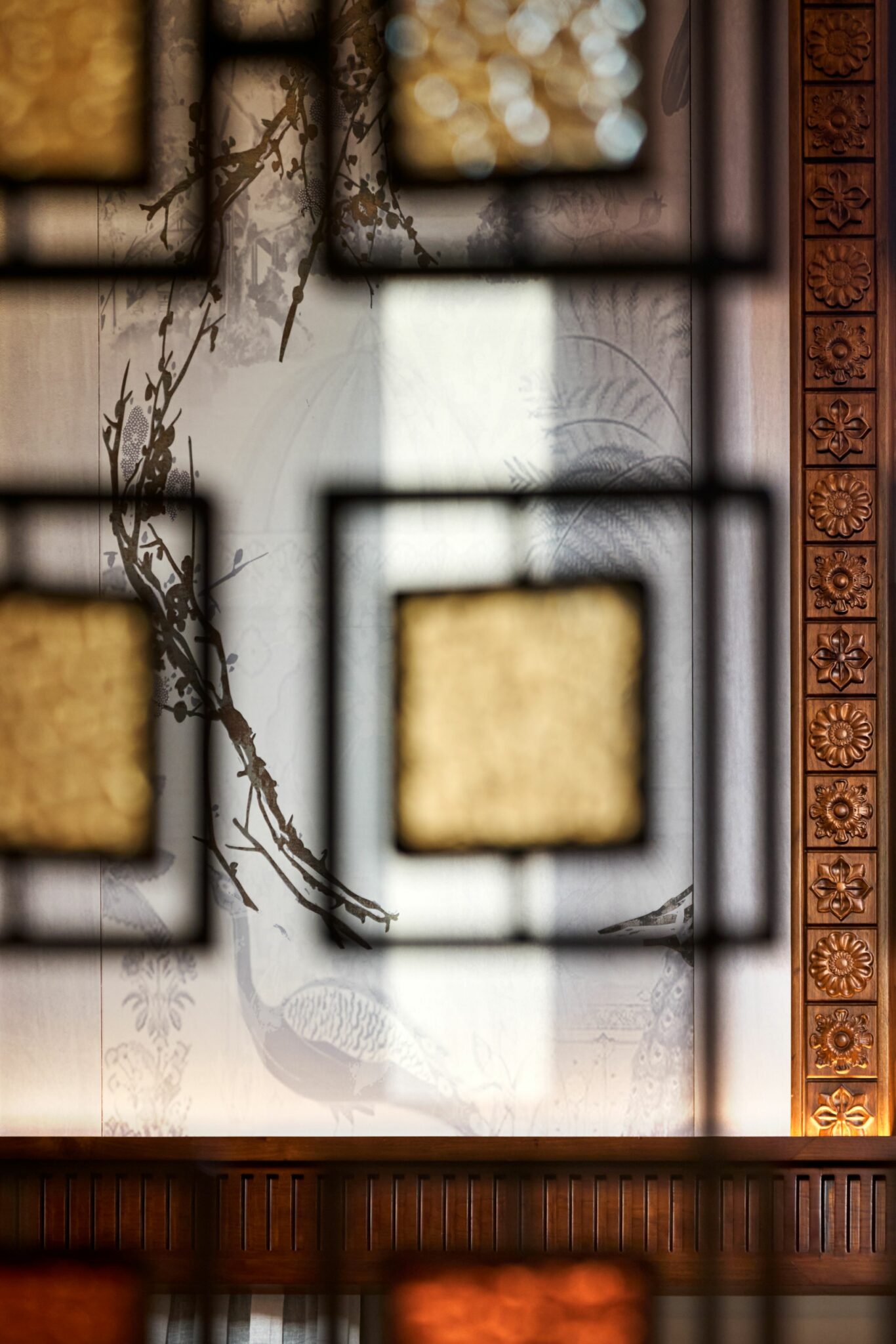 Ornate room divider with square panels in focus. Blurred mural of branches and peacock behind. Right side features a carved wooden wall panel. Warm tones.
