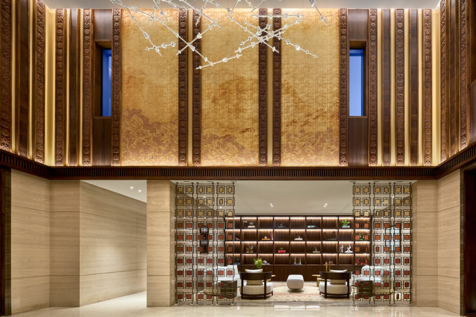 Luxurious hotel lobby with high ceilings adorned with ornate wood panels and warm lighting. Modern seating, decorative screens, and elegant chandelier.