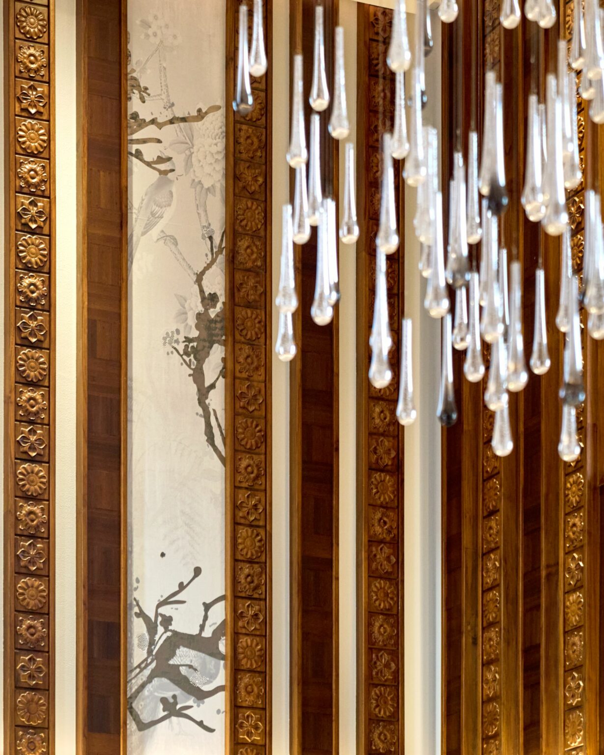 Ornate wood paneling with floral carvings and a delicate nature mural create an elegant backdrop. Nearby, a modern chandelier with tear-shaped glass accents adds a contemporary touch.