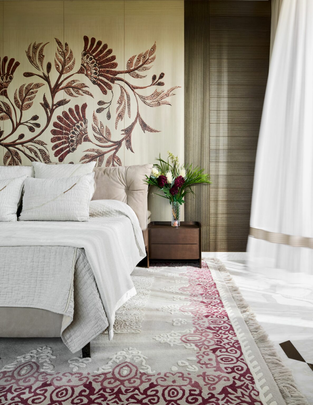 A cozy bedroom features a beige bed with white pillows and linens, a floral wall mural, and a nightstand with a vase of flowers. A patterned rug adds color and warmth.