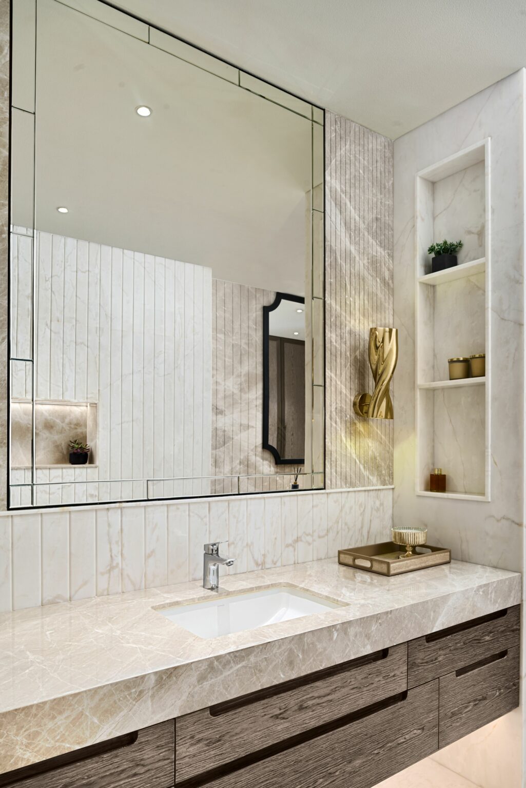 Modern bathroom with a large marble countertop, sleek faucet, and decorative items. Features a broad mirror, geometric wall light, and small potted plant.