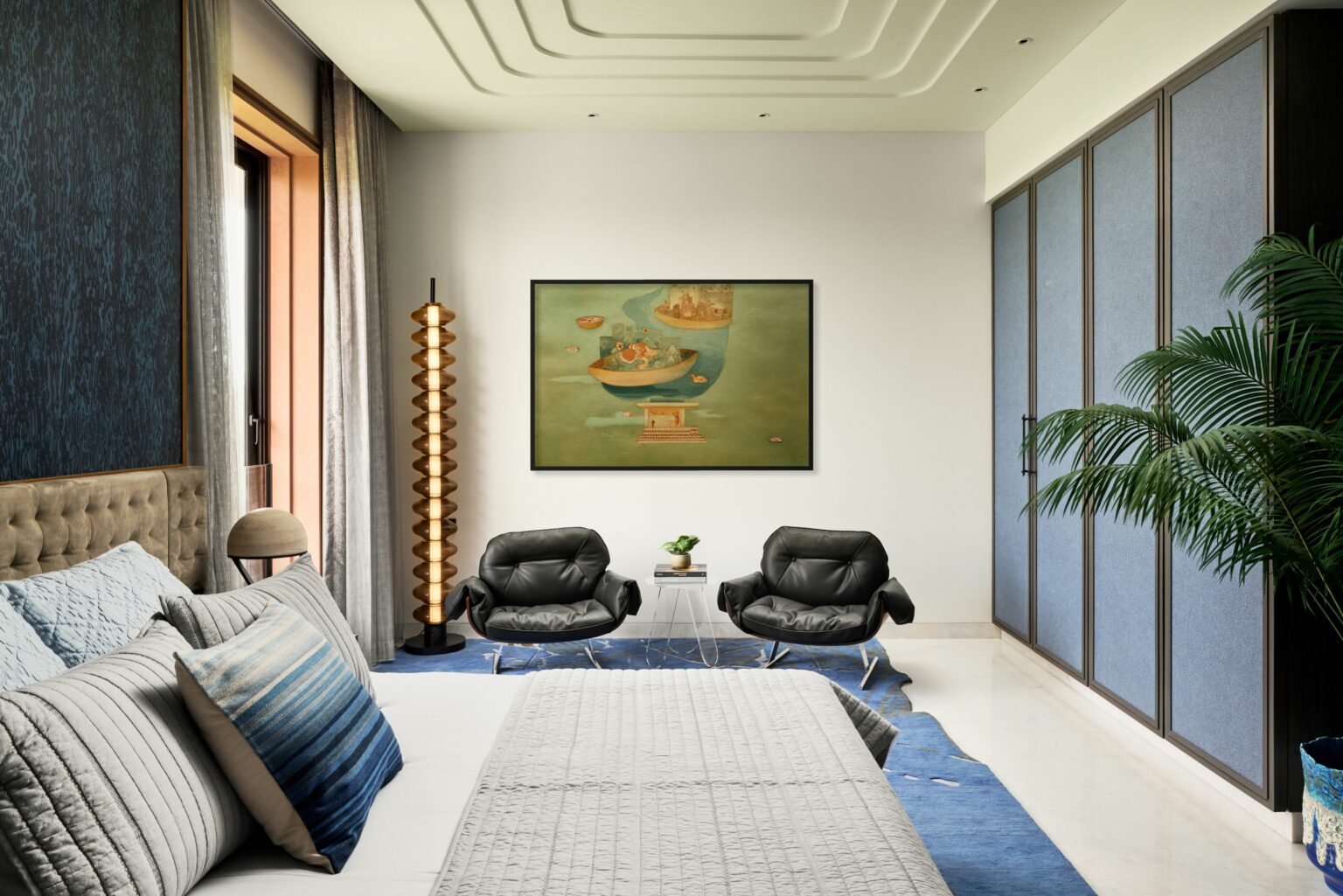 Modern bedroom with a large bed featuring blue and gray pillows, two black armchairs, a tropical plant, artistic wall painting, and a geometric lamp. Calm ambiance.