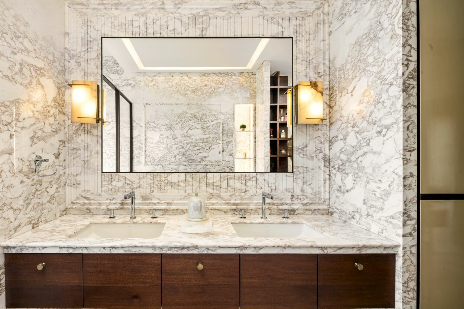 Luxurious marble bathroom with dual sinks, under large mirror. Warm lighting sconces, wood cabinetry, and glass cloche create an elegant, serene mood.
