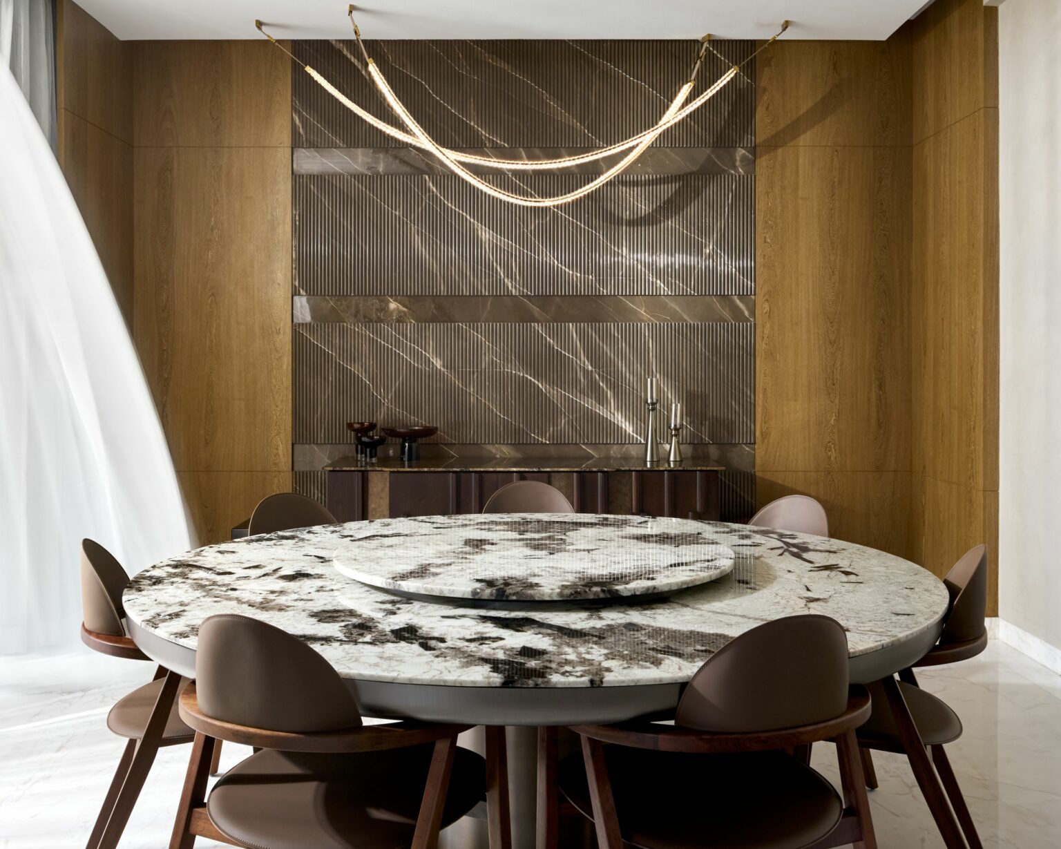 Elegant dining room with a round marble table, brown chairs, and sleek wood paneling. A flowing curtain and artistic lighting add a sophisticated touch.