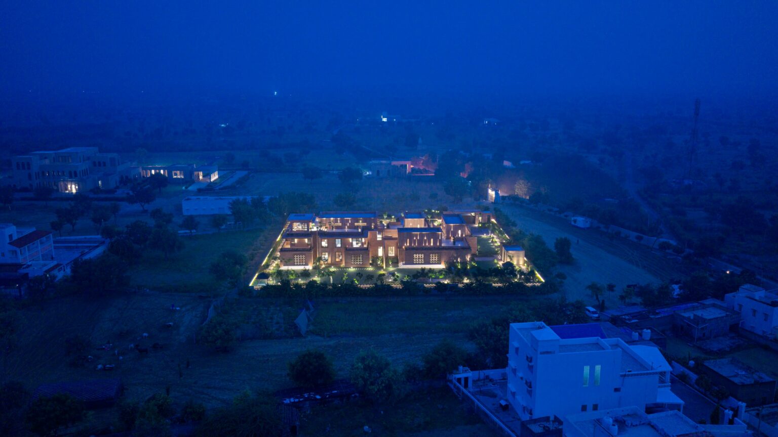 Aerial view of a large, illuminated building surrounded by greenery at night. The building emits a warm glow, contrasted by the deep blue of the evening sky.