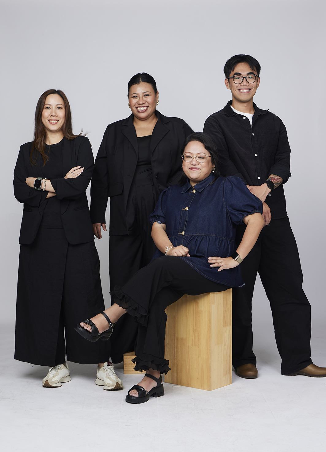 A group portrait of four individuals smiling confidently against a gray backdrop. Three are standing and one is seated on a wooden stool, conveying teamwork and positivity.