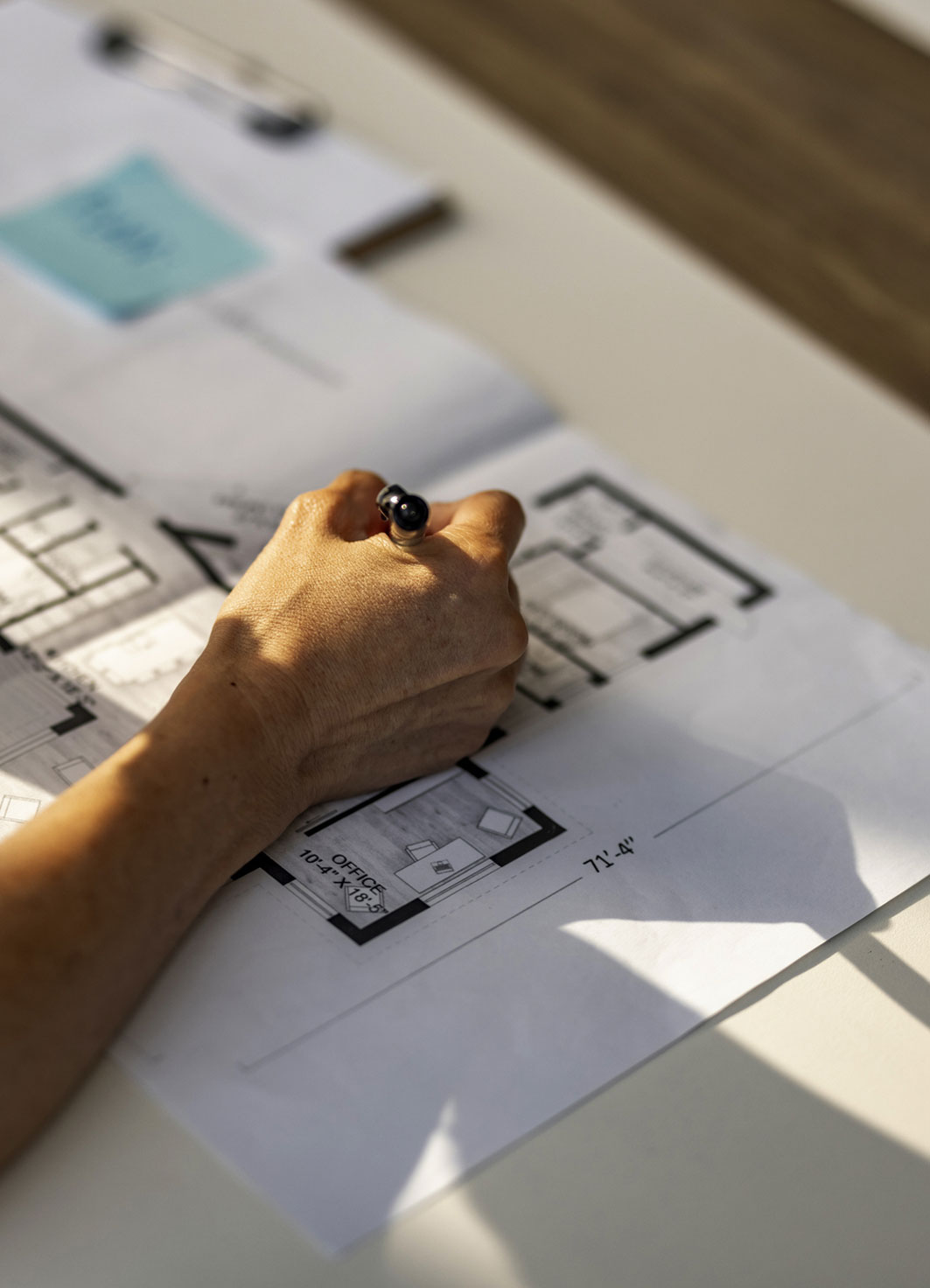 A hand holds a pen, poised over a detailed architectural floor plan on a table, suggesting focus and planning. Natural light casts soft shadows.