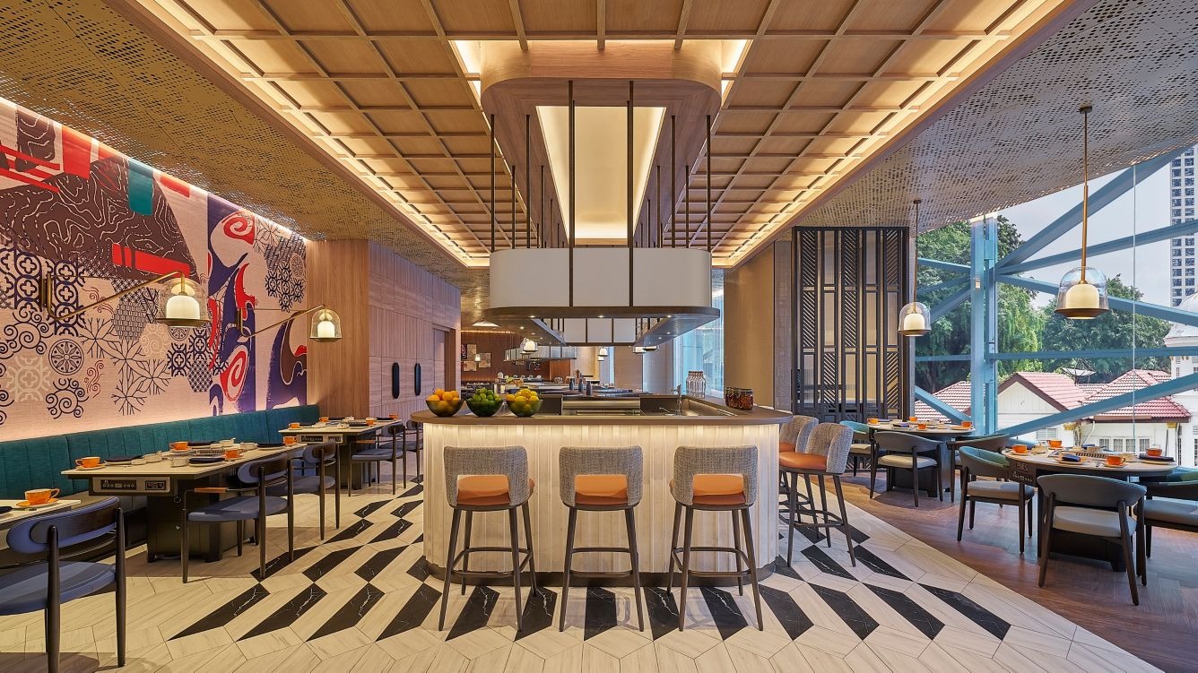 Modern restaurant interior with a central bar, stylish wooden ceiling, and patterned floor. Vibrant mural on the left wall, cozy seating on both sides, and large windows reveal trees outside.