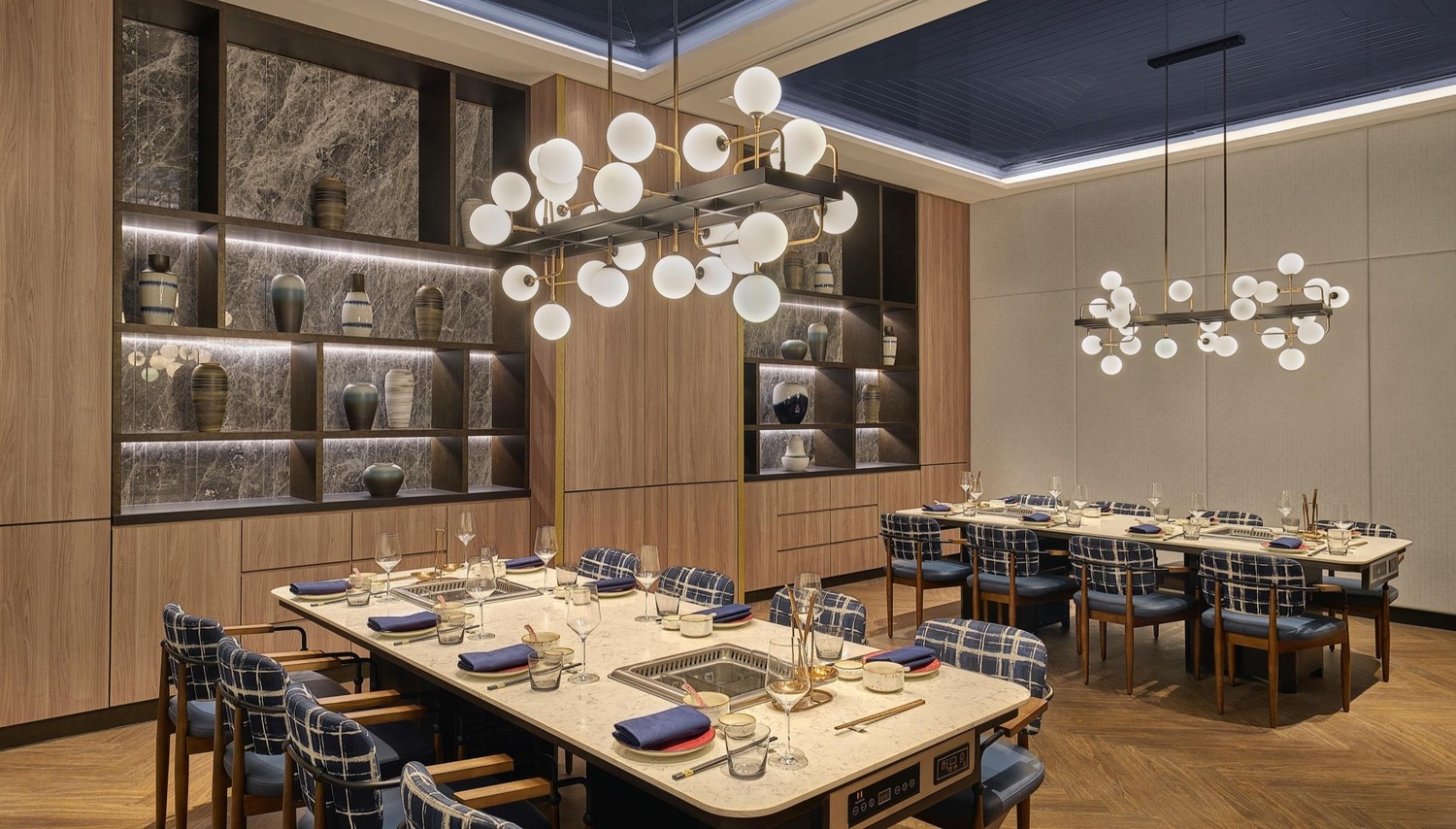 Spacious dining room with two tables set for dining, surrounded by blue plaid chairs. Modern chandeliers hang overhead. Elegant, warm ambiance.