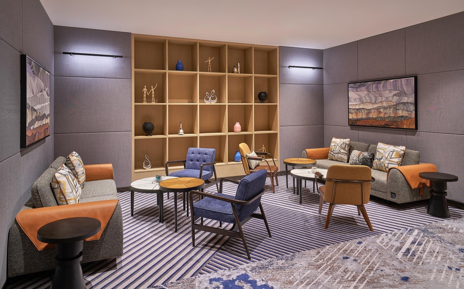 A stylish lounge features modern gray sofas adorned with orange throws, blue and beige armchairs, circular tables, and a wooden shelf with decorative items.