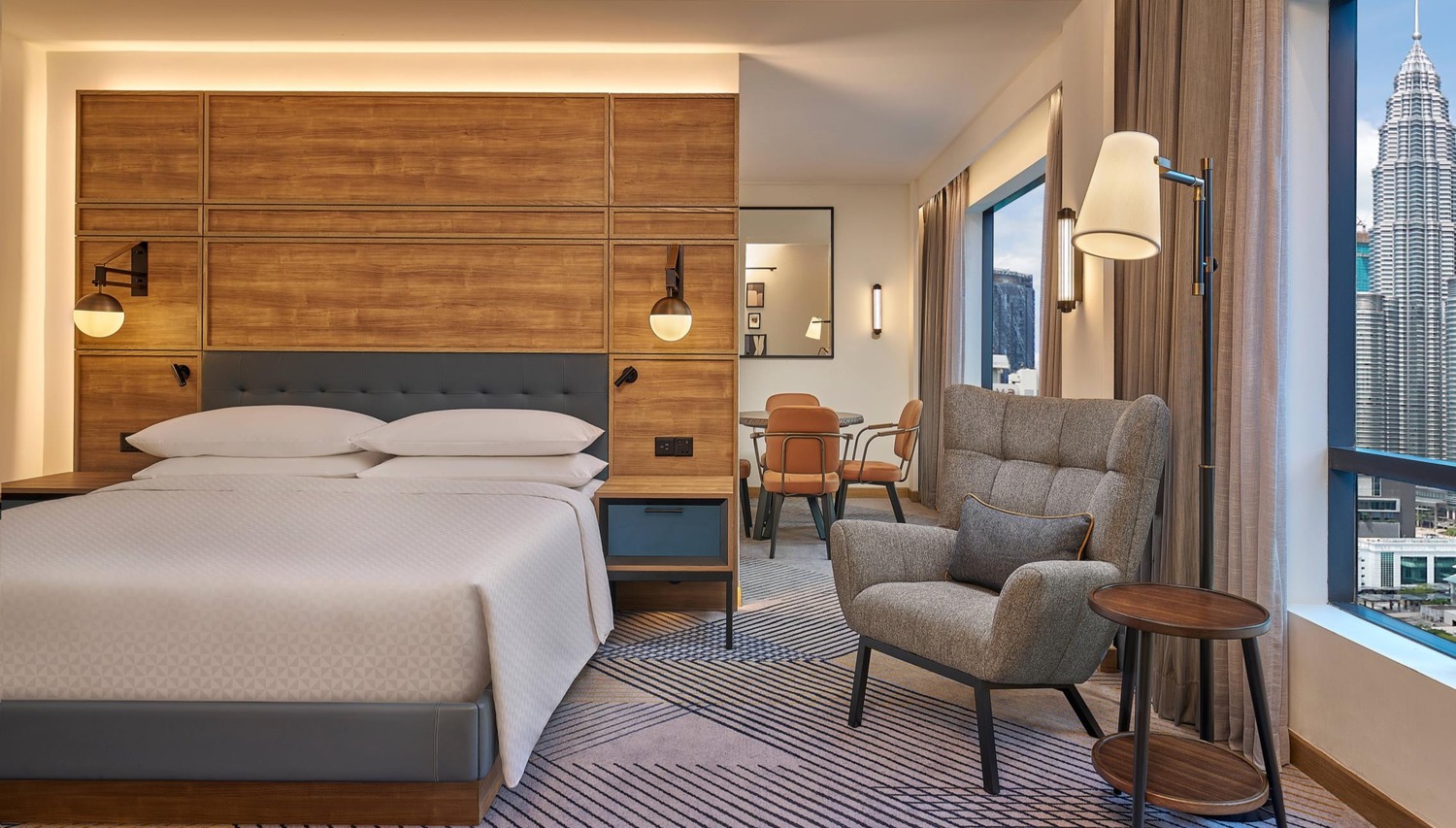 Modern hotel room with cozy lighting, featuring a large bed, cushioned armchair, and a view of city skyscrapers through large windows. Elegant and inviting.