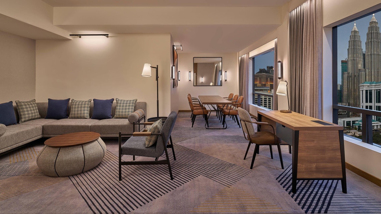 Sophisticated hotel suite with a modern lounge area, dining table, and desk by large windows revealing a cityscape, including two skyscrapers.