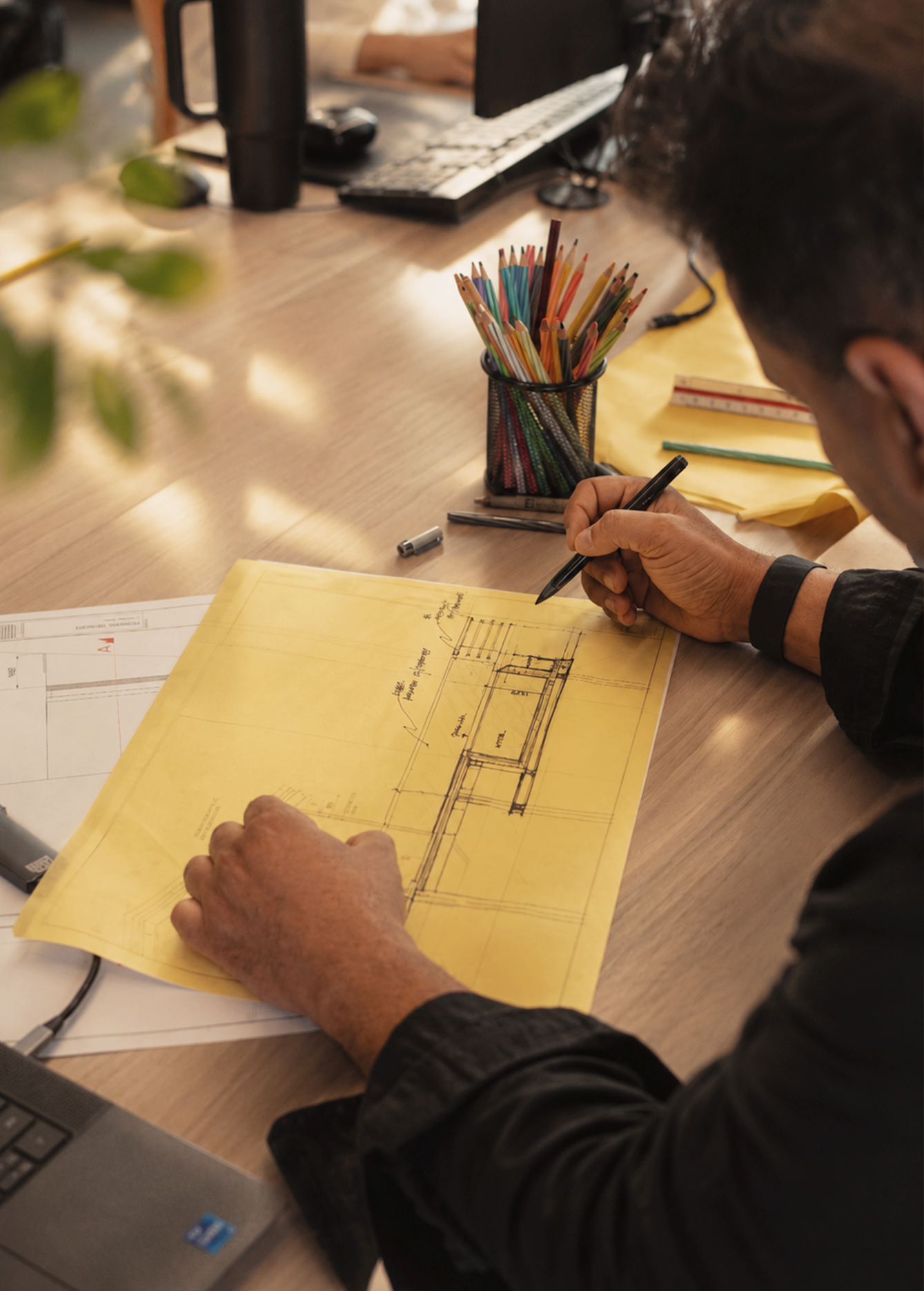 A person is sketching architectural plans on yellow paper at a desk with colored pencils and a laptop. The scene conveys focus and creativity.