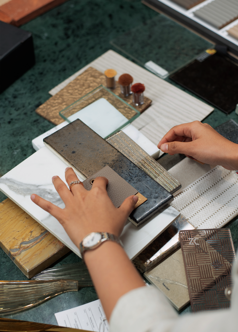 A person's hands arrange various materials like wood, metal, and fabric samples on a green table, suggesting a design selection process.