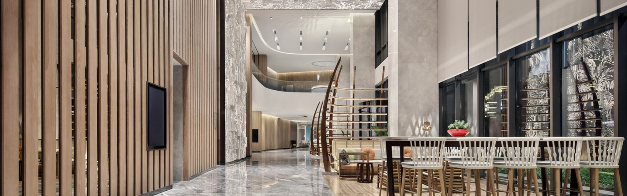 Spacious modern lobby with marble floors, high ceilings, and stylish wood accents. A curved staircase leads up, and tall windows line the right side.