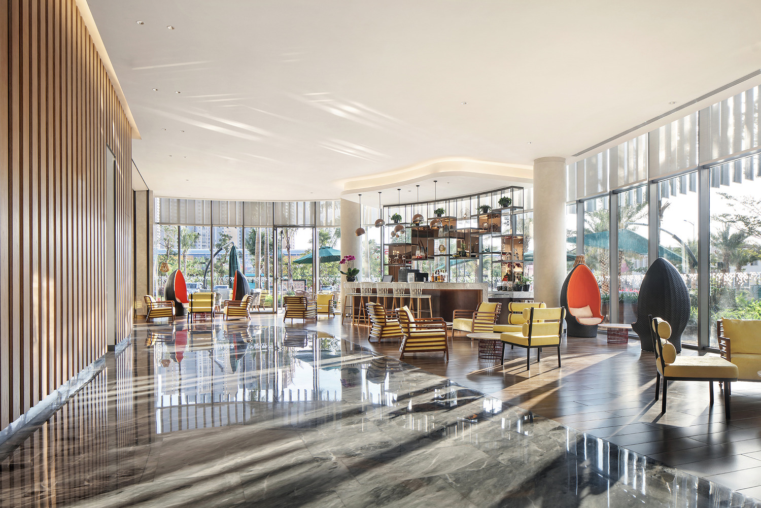 Spacious, modern hotel lobby with floor-to-ceiling windows. Sunlit seating area features striped chairs, hanging egg chairs, and a central bar. Bright, airy atmosphere.